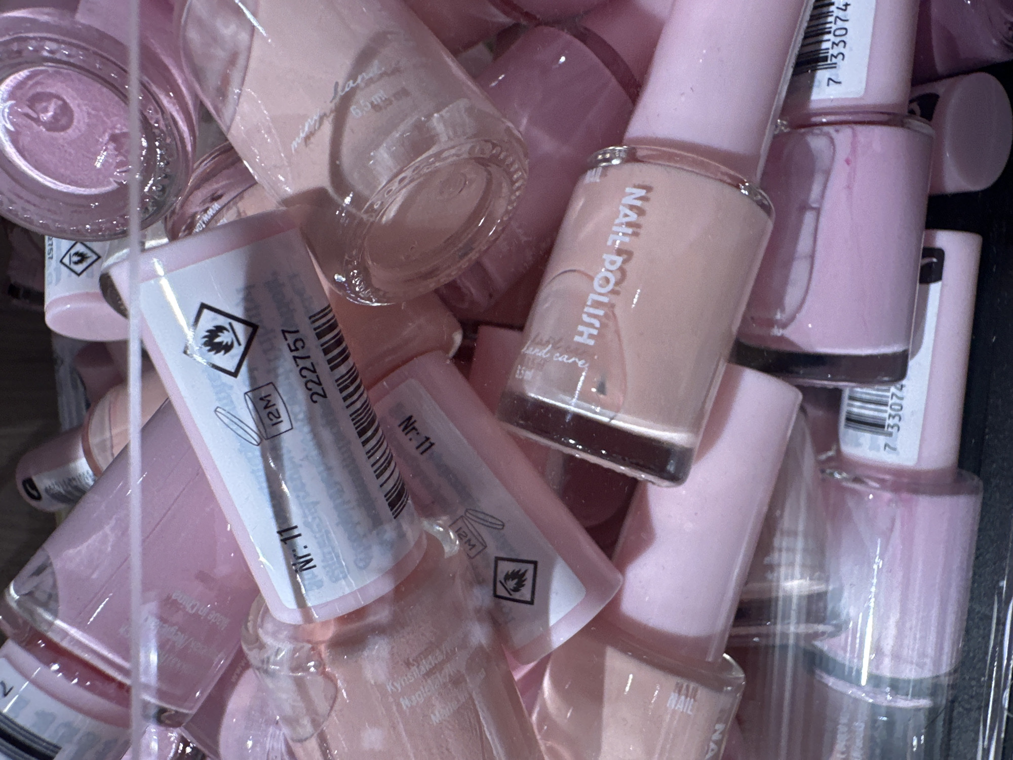 The image displays a close-up, overhead view of a large collection of pink nail polish bottles. The bottles are packed tightly together, with some appearing to be stacked on top of others. Several bottles have visible labels. One label shows "Nr. 11" and a "12M" symbol, indicating shelf life. Another label has a symbol of a flame within a triangle, signifying flammability. A partially visible bottle in the foreground has the words "NAIL POLISH" printed vertically. The lighting is bright, casting reflections on the glass bottles. The overall scene appears to be a retail display or storage area for cosmetics.