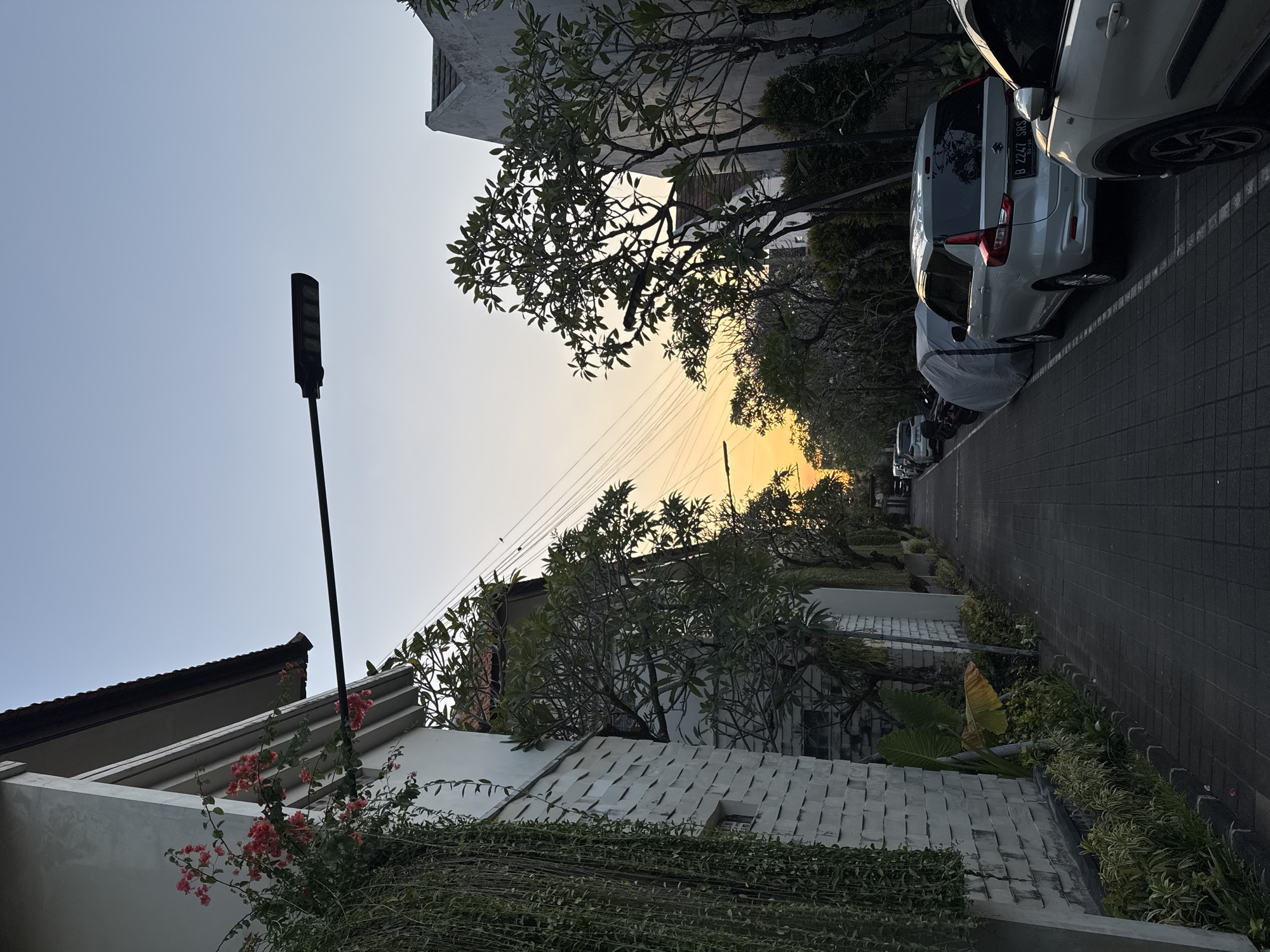 This image depicts a street scene in Denpasar, Indonesia, during sunset. A narrow paved street runs into the distance, lined with parked cars and houses. On the left, a white house with a tiled roof features a white textured wall and hanging plants. Red flowers bloom on a vine cascading down the side of the house. Across the street, lush green trees and more houses are visible. The sky displays a gradient of colors, from a pale blue at the top to a warm orange and yellow near the horizon, indicating the setting sun. A modern street lamp extends over the roadway. Several cars are parked along the right side of the street, with one white SUV prominently featured. The atmosphere is calm and quiet, suggesting a residential area.