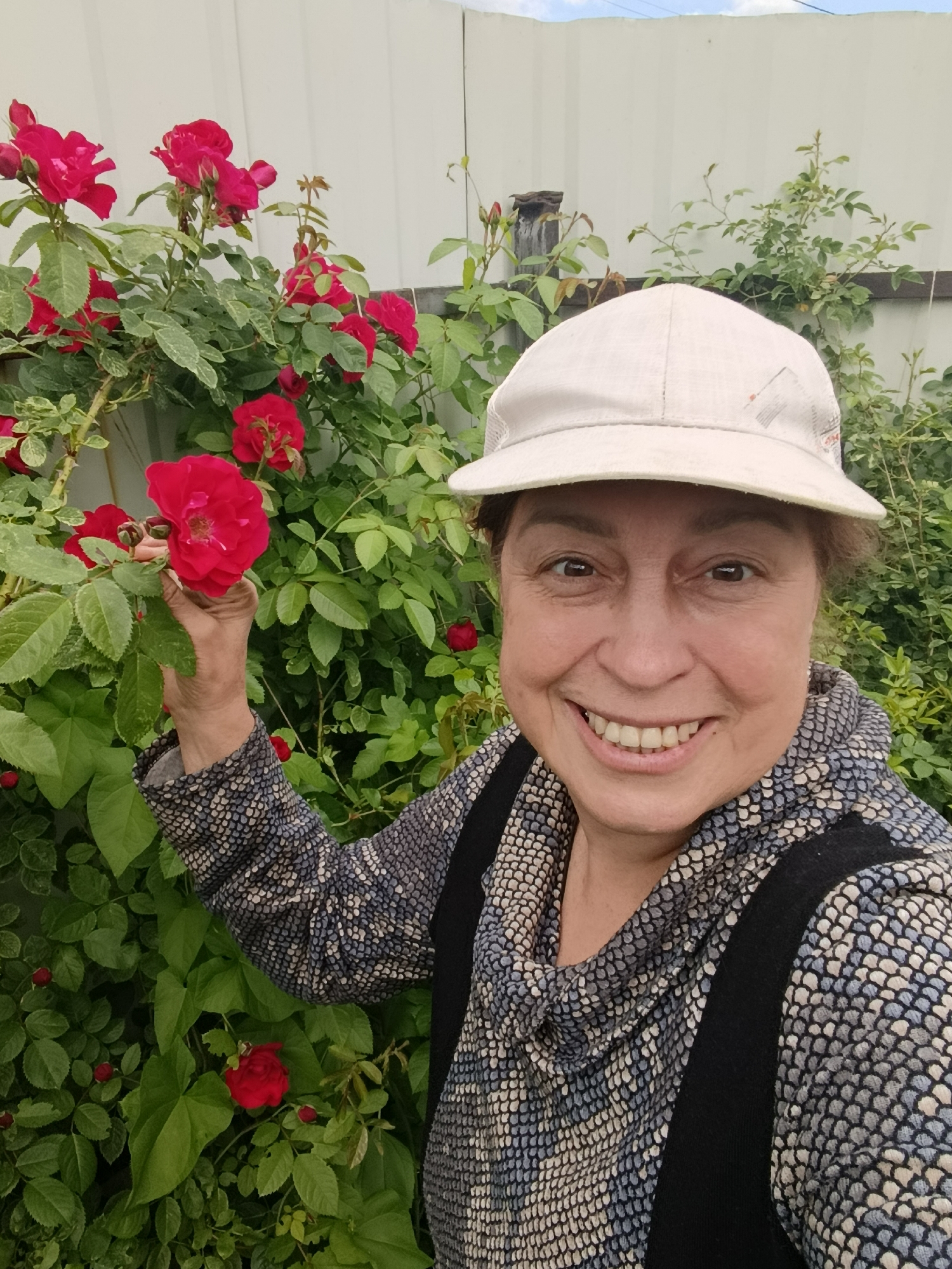 A middle-aged woman wearing a white baseball cap and a patterned long-sleeved shirt is posing for a selfie in front of a rose bush. She is smiling and holding a red rose. The background consists of green foliage, a white fence, and a partly cloudy sky. The scene appears to be an outdoor garden.