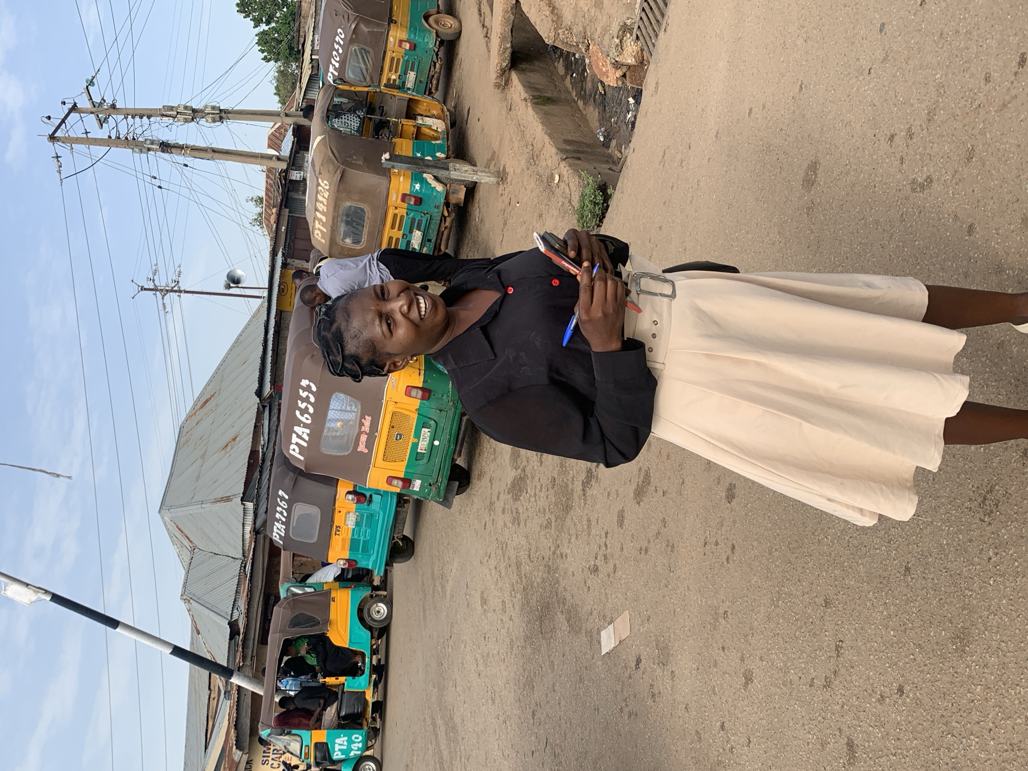 This is a vertical, low-angle, full-body shot of a Black woman standing on a dusty street in Gura Top, Nigeria, with a line of colorful auto-rickshaws parked behind her. The woman is smiling at the camera and holding a red pen and a black phone. She is wearing a black, collared shirt and a light-colored, knee-length skirt with a wide, silver buckle belt. The auto-rickshaws are a mix of yellow, green, and brown. Some have text visible, including "PTA-7367", "PT-10570-", "PTA-6553", "PT-14126", and "PTA-740". There are power lines and poles overhead, suggesting an urban or semi-urban environment. The ground is a rough, textured asphalt. The lighting suggests it is daytime. The woman's expression and the bright colors of the rickshaws contribute to a lively and cheerful atmosphere.