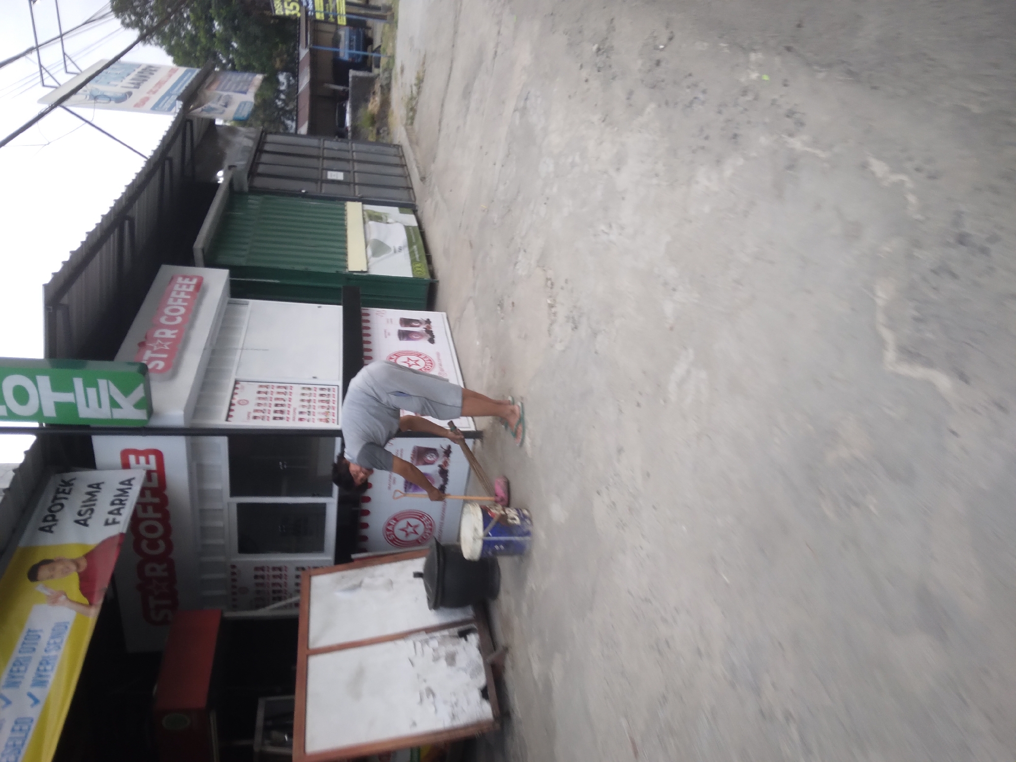 In Desa Pamijen, Kecamatan Sokaraja, Indonesia, a person is sweeping the pavement in front of a small business. The individual, dressed in a grey t-shirt and shorts, is using a broom and a pink dustpan, with a blue bucket nearby. The business appears to be a "Star Coffee" establishment, with signage visible on its facade. The surrounding area shows a typical street scene, with a paved road and a sky that suggests an overcast day. The overall impression is one of routine daily activity.