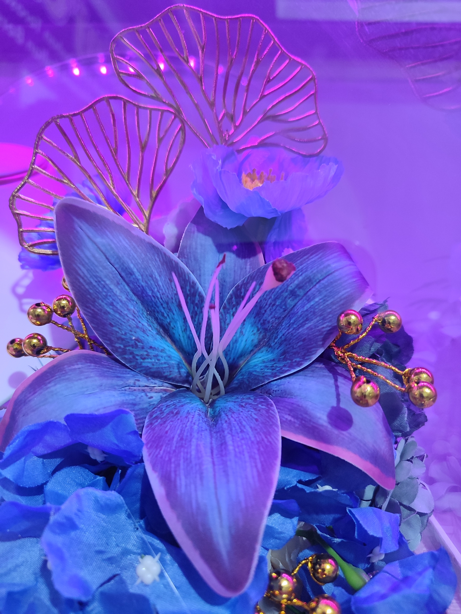 The image shows a close-up of an artificial flower arrangement. The prominent flower is a large, deep blue lily with iridescent purple and teal markings on its petals. The center of the lily features pink stamen and pistil. Surrounding the lily are smaller blue flowers and blue fabric petals. Decorative elements include golden wire accents with small gold bead-like spheres. In the background, there are two stylized gold leaf-like embellishments and another purple flower with a yellow center. The entire scene is bathed in a strong purple light, creating a surreal and vibrant atmosphere. There are no people or specific locations visible that would allow for identification of Gantungan, Indonesia.