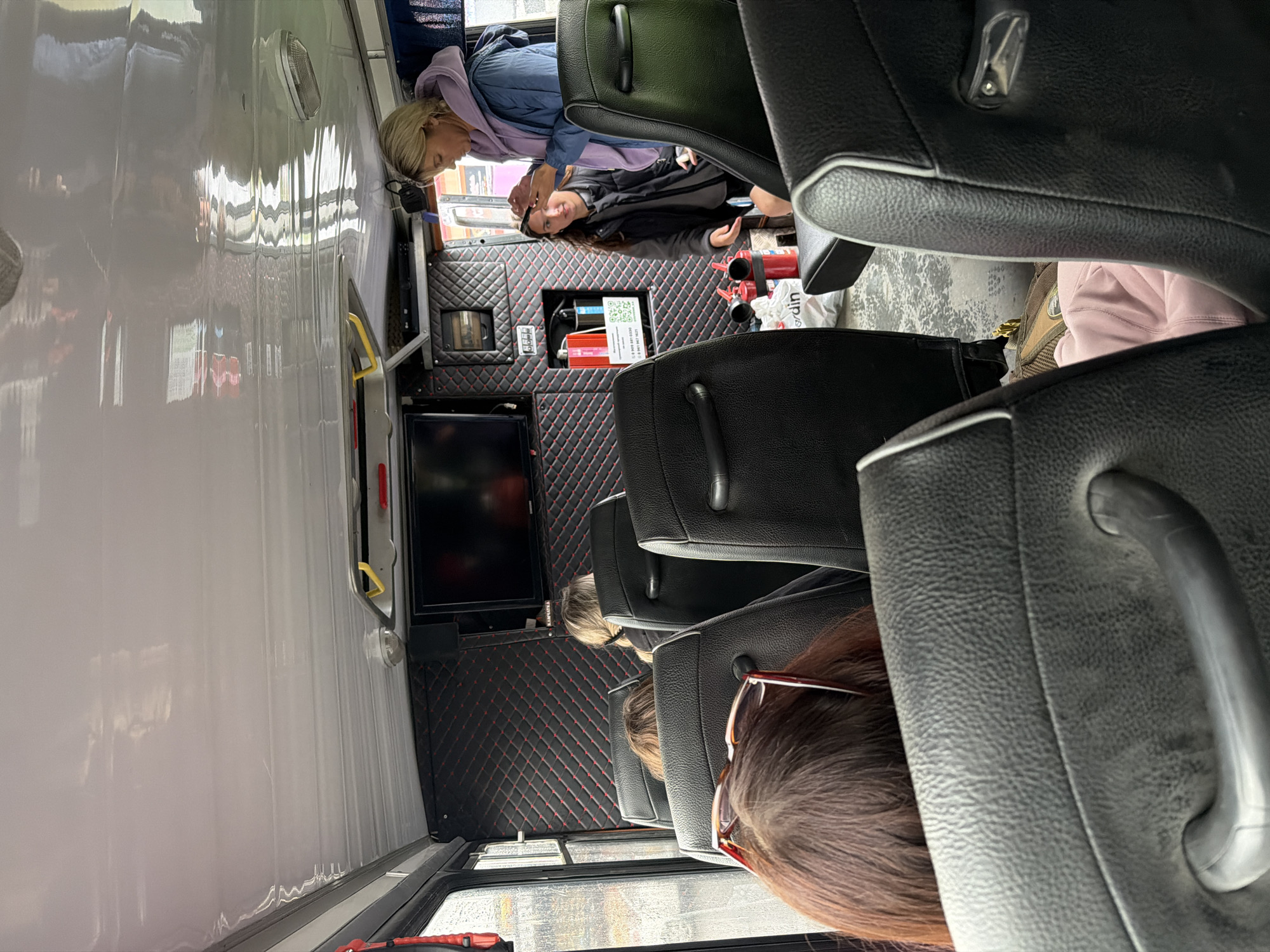 Inside a vehicle, possibly a bus or van, passengers are seated. In the foreground, a person's head and shoulders are visible, with sunglasses resting on their hair. In the mid-ground, a woman with blonde hair, wearing a blue jacket and a purple hoodie, stands and appears to be interacting with another passenger, a person with long dark hair wearing a dark vest. The interior of the vehicle features black seats with silver trim and a black quilted wall panel with red stitching. A television screen is mounted on the wall. Red fire extinguishers are visible on the floor. The scene appears to be located in Petropavlovsk-Kamchatsky, Russia, although no specific landmarks are identifiable in the image.