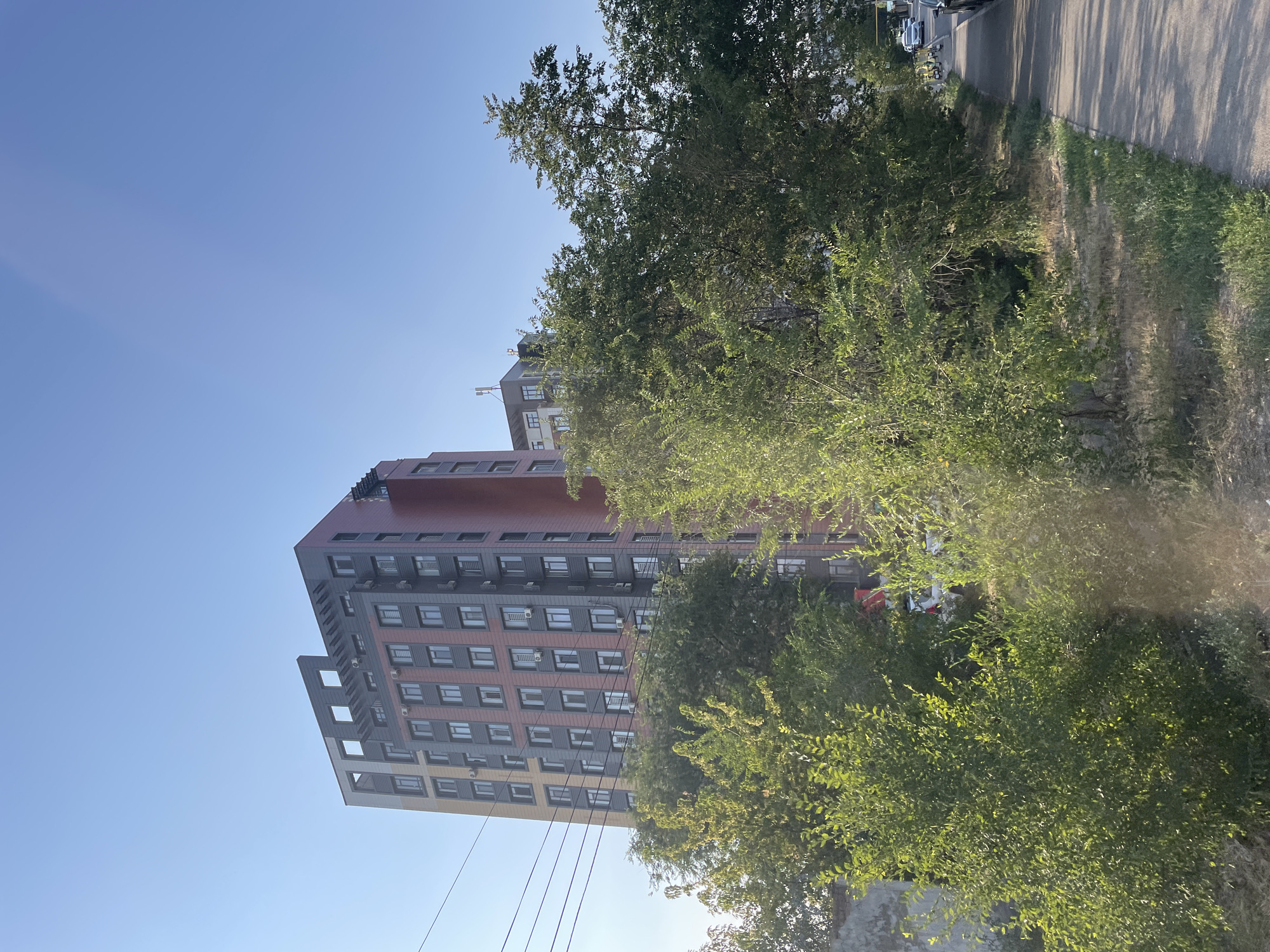 A modern, multi-story apartment building with a facade of gray, red, and tan panels is visible in the city of Almaty, Kazakhstan. The building has numerous windows and visible air conditioning units. In front of the building, there are several large, green trees. A paved sidewalk runs along the right side of the image, adjacent to a grassy area and road. The sky is clear and blue.