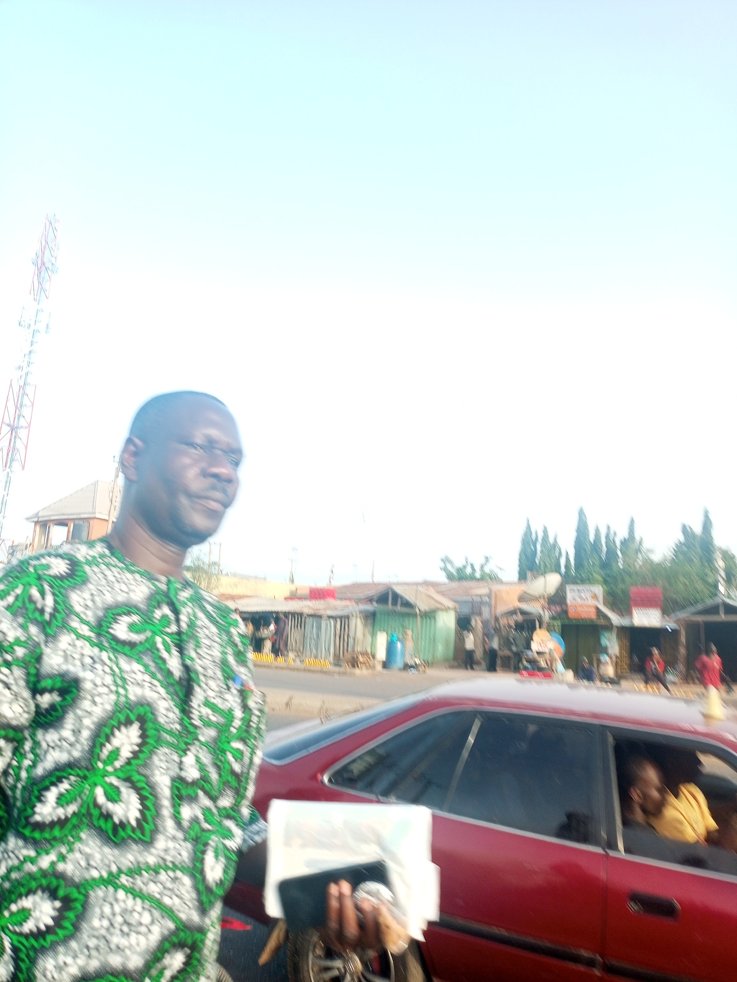 A man in a green and white patterned shirt stands near a red car with a person inside, likely in the city of Tumfure, Nigeria. A cell tower is visible in the background, along with buildings and stalls. Other people are also present in the background. The man is holding something, appearing to be a phone, in his hands.