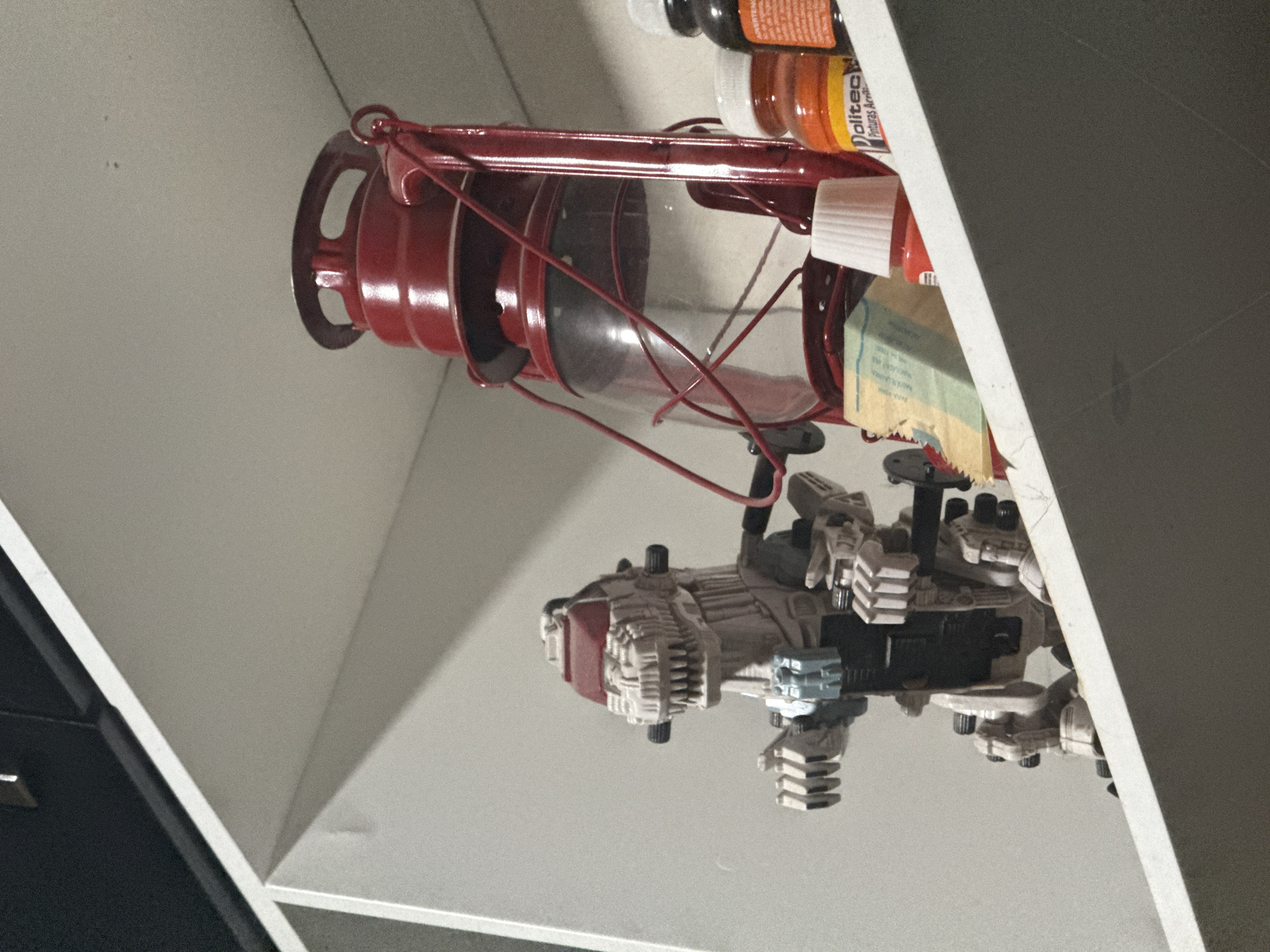 The image, taken in Puebla, Mexico, depicts an indoor still life scene featuring various objects arranged on a shelf or within a cubby. The frame is dominated by a red, vintage-style lantern with a clear glass globe on the left. Below and to the right of the lantern is a detailed model or toy, possibly a futuristic vehicle or robot, predominantly white and light grey with black and red accents, displaying intricate mechanical parts.
Behind the lantern, on what appears to be a shelf, are several small bottles and jars. One bottle clearly shows the text "Dolitect Pinturas Acrílicas," indicating it contains acrylic paints. Other bottles, with white or black caps, hold various colored liquids, likely also craft supplies. A crumpled, light brown paper bag or small box with light blue markings is positioned below these paint bottles.
The scene is framed by white and dark grey structural elements, suggesting the objects are housed within a shelving unit or a personal display area. No activity is taking place, and the lighting is even, illuminating the items in a clear, natural way. The arrangement suggests a personal space, possibly a craft area or a collector's display.