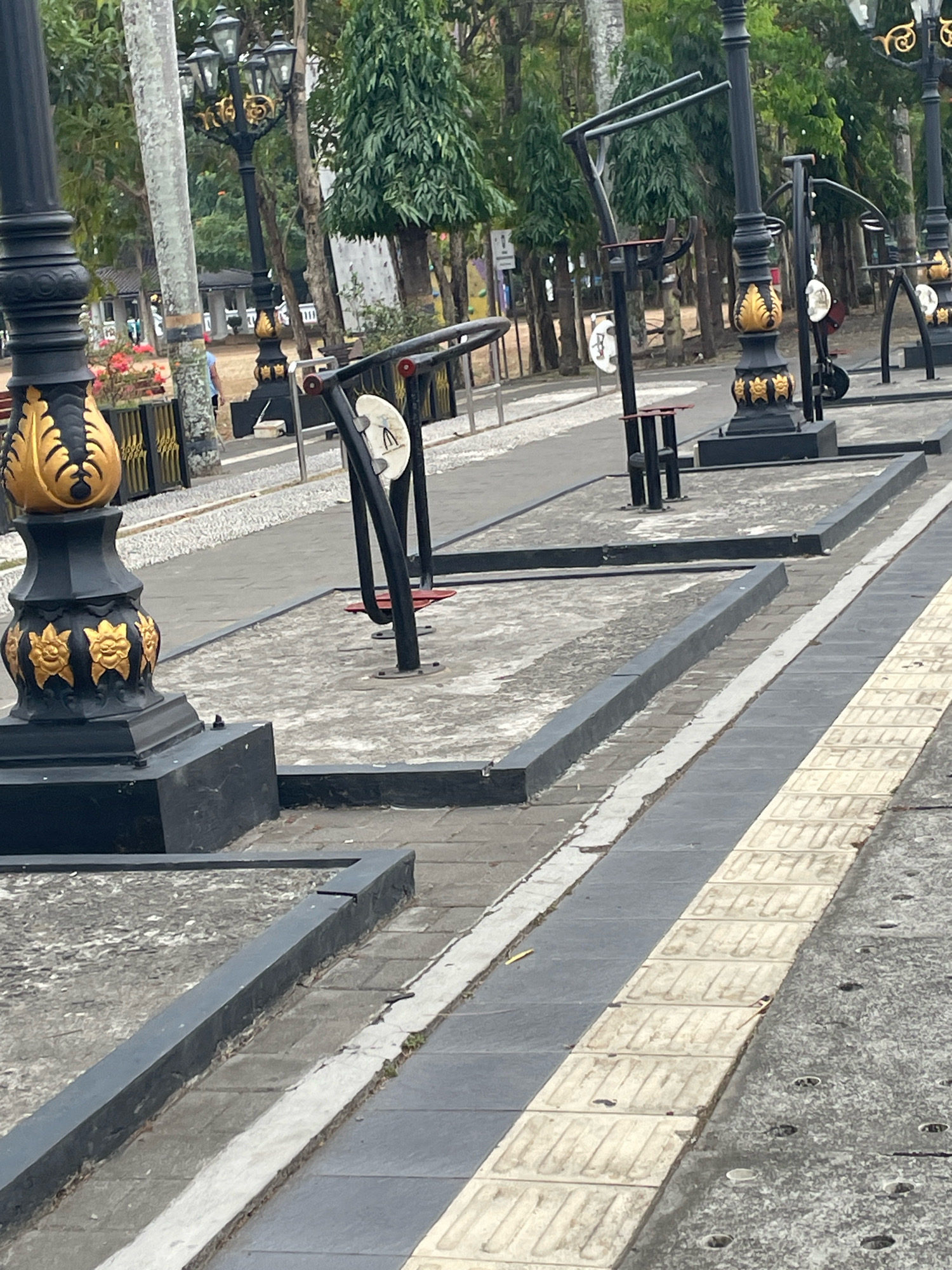 The image shows an outdoor exercise equipment area in the city of Purworejo, Indonesia. The equipment consists of metal frames designed for various exercises. In the foreground, there is a decorative black lamppost with gold accents. The ground is paved with gray and white tiles. In the background, there are trees and a white building. The scene appears to be a public park or recreational area.