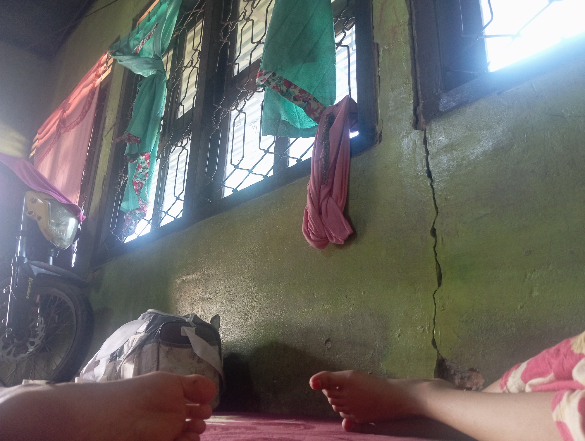 The image depicts an indoor scene, likely in Rejosari, Indonesia. The focus is on a wall painted green, which is cracked vertically in the center. To the left of the wall, a motorcycle is partially visible, with its headlight and front wheel in view. A bag or sack with a grey and white patterned design rests on the floor next to the motorcycle. Two feet are prominently positioned in the foreground, indicating the presence of at least one person. Several pieces of fabric, appearing to be clothing or curtains, are hanging from a barred window on the upper left of the image. One piece is pink and draped over the motorcycle's headlight, another is teal with floral patterns hanging in front of the window, and a pink piece is also hanging near the window. The overall environment suggests a domestic or storage space. The lighting is harsh, with a strong source of light coming from the right side of the window, suggesting natural daylight entering the room.