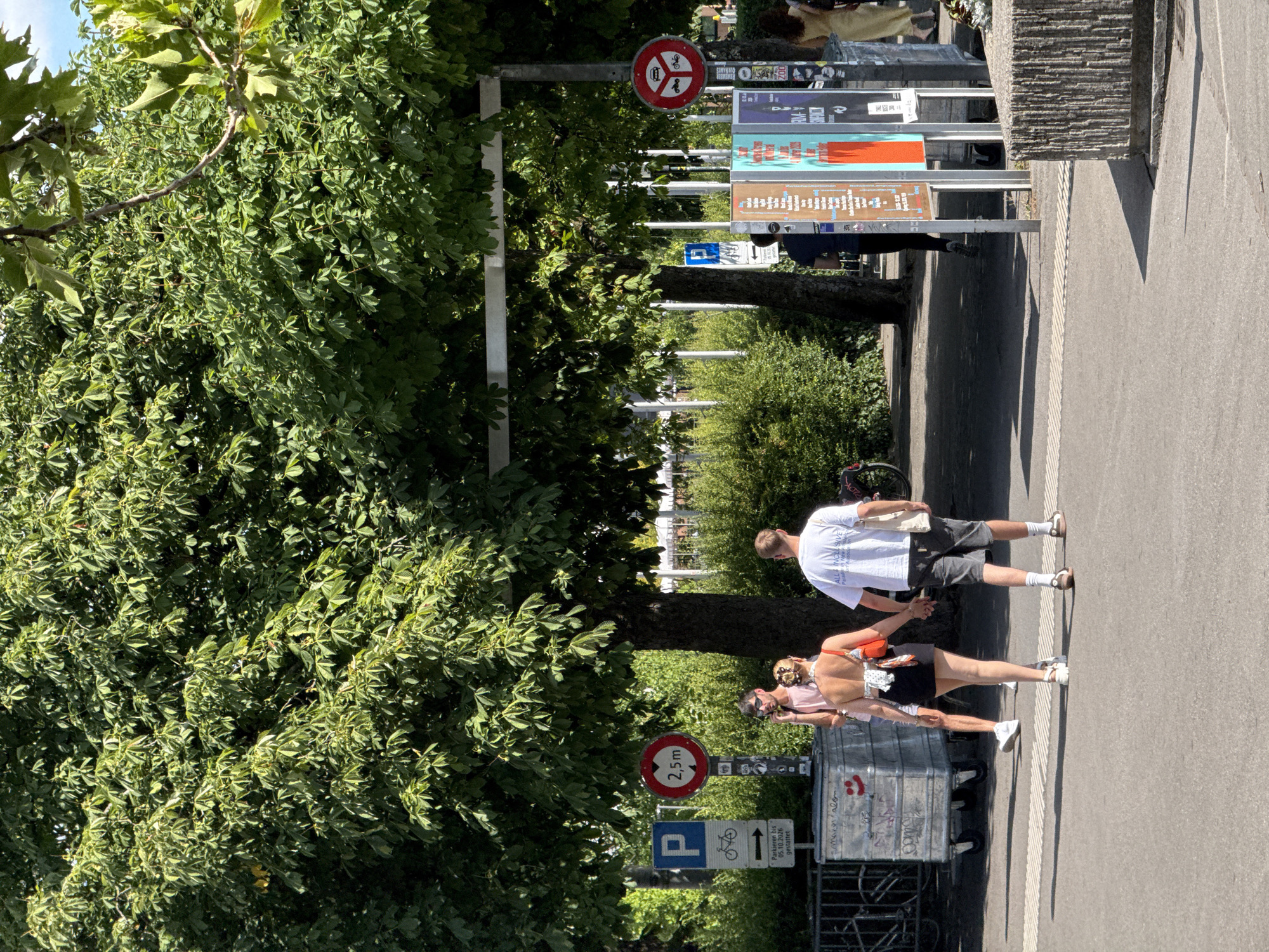 The image depicts a scene in Zurich, Switzerland, likely near a park or public space, characterized by lush green trees, paved walkways, and urban elements. In the foreground, a couple is walking hand-in-hand across a crosswalk. The woman is wearing a white patterned bandeau top, a black mini skirt, and white sneakers, while the man is in a white t-shirt, dark shorts, and white socks with brown sneakers. They are walking away from the camera.
Behind them, a silver industrial bin with graffiti is visible on the left. Several traffic and parking signs are posted on poles. One sign indicates "P" for parking with a bicycle symbol and an arrow, and text reading "Parkieren bis 05.10.2026 gestattet." Another circular sign shows a red border with a diagonal line and "2,5m" indicating a height restriction.
Further back, a group of people are walking. One individual is talking on a cell phone, wearing a pink t-shirt and sunglasses. A series of colorful display boards are mounted on poles, featuring various posters with text, some of which appear to be event advertisements. The environment is sunny, with shadows cast on the ground, suggesting it is daytime. The background shows more greenery, possibly bushes and trees, and some white structures that might be part of urban infrastructure. A woman in a yellow dress is also visible in the far right of the image.