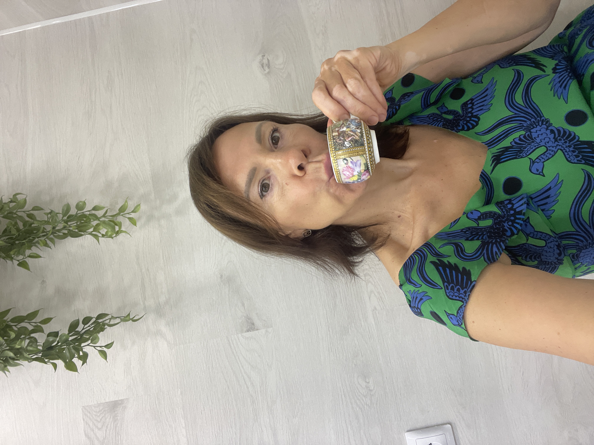 A medium shot shows a woman with medium brown hair, wearing a green dress with a blue bird pattern, holding a small, ornate teacup to her lips, as if drinking. She is positioned in the center of the frame, against a backdrop of a pale wood-paneled wall and a white electrical outlet on the left. Two hanging artificial green plants are visible in the upper left quadrant of the image. The woman is looking towards the camera. The scene appears to be indoors, possibly in a domestic setting. There are no identifiable landmarks or features that would specifically place the location in Sochi, Russia.
