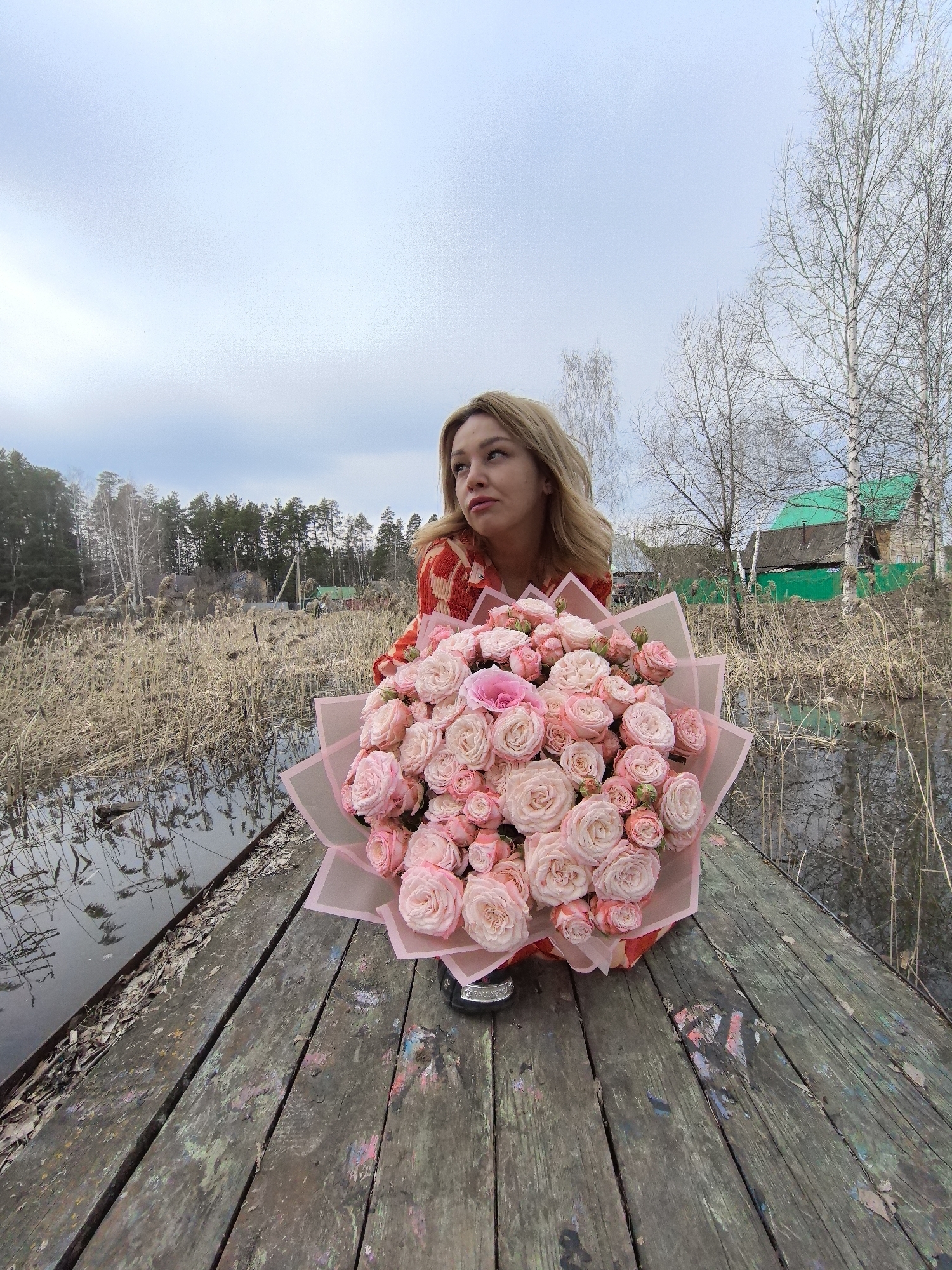 A young woman with blonde hair is sitting on a weathered wooden surface, possibly a dock or platform, and holding a large bouquet of pink roses. The setting appears to be near a body of water, with reeds and vegetation visible in the background, and a glimpse of a green-painted building. The sky is overcast. The overall scene evokes a tranquil, perhaps melancholic, atmosphere. This is not necessarily Noviy, Russia, unless corroborated by additional data.