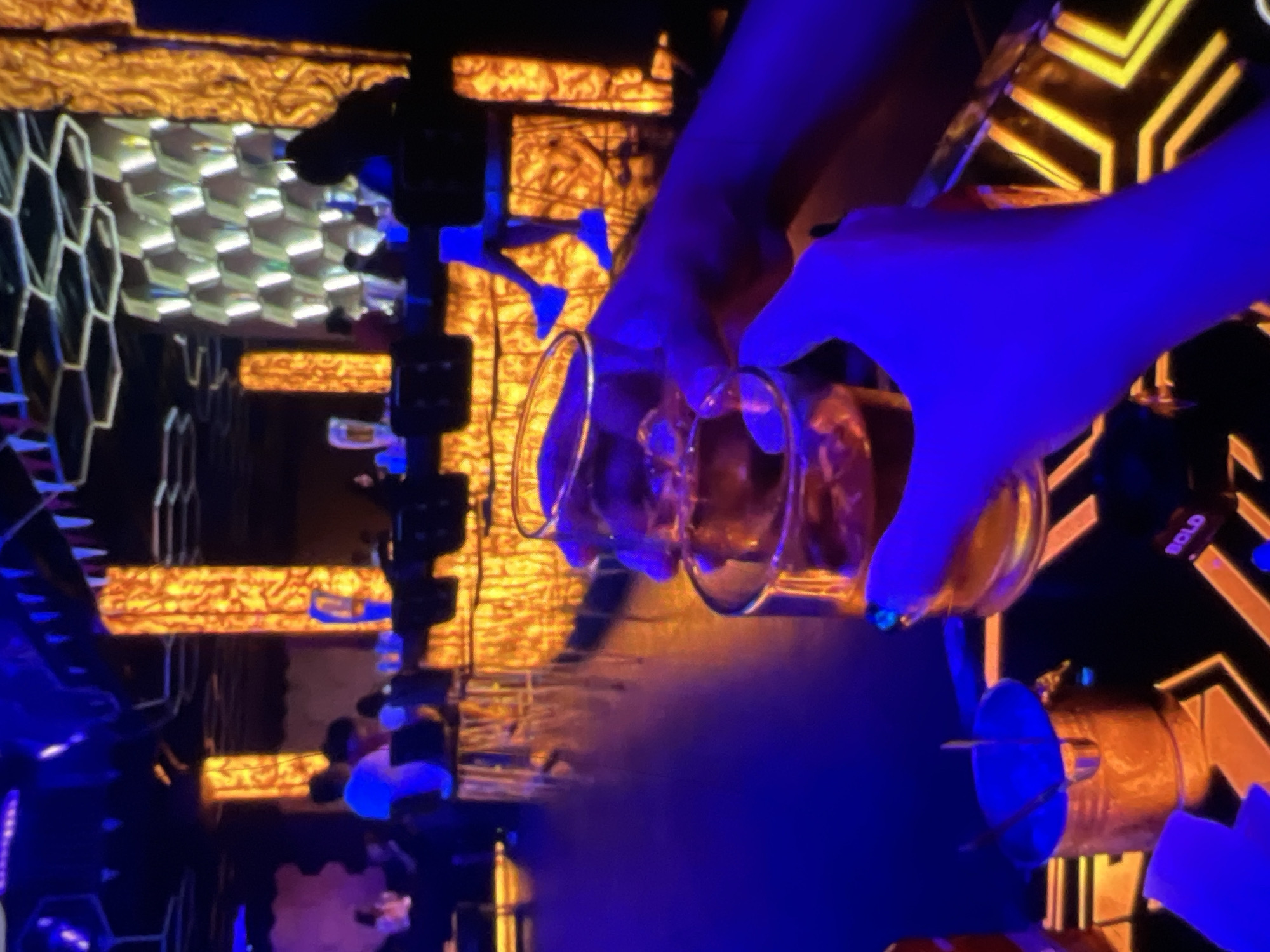 The image captures a dimly lit, vibrant bar scene in Lagos, Nigeria, characterized by intense blue and orange lighting. In the foreground, two hands are clinking glasses filled with amber liquid, likely alcoholic beverages, over a black and orange patterned table. An ice bucket, appearing to be made of hammered gold-colored metal, is positioned to the left, holding ice tongs. In the background, patrons are seated at a bar, illuminated by glowing orange pillars and a hexagonal wall feature. The overall atmosphere suggests a lively and upscale nightlife environment.