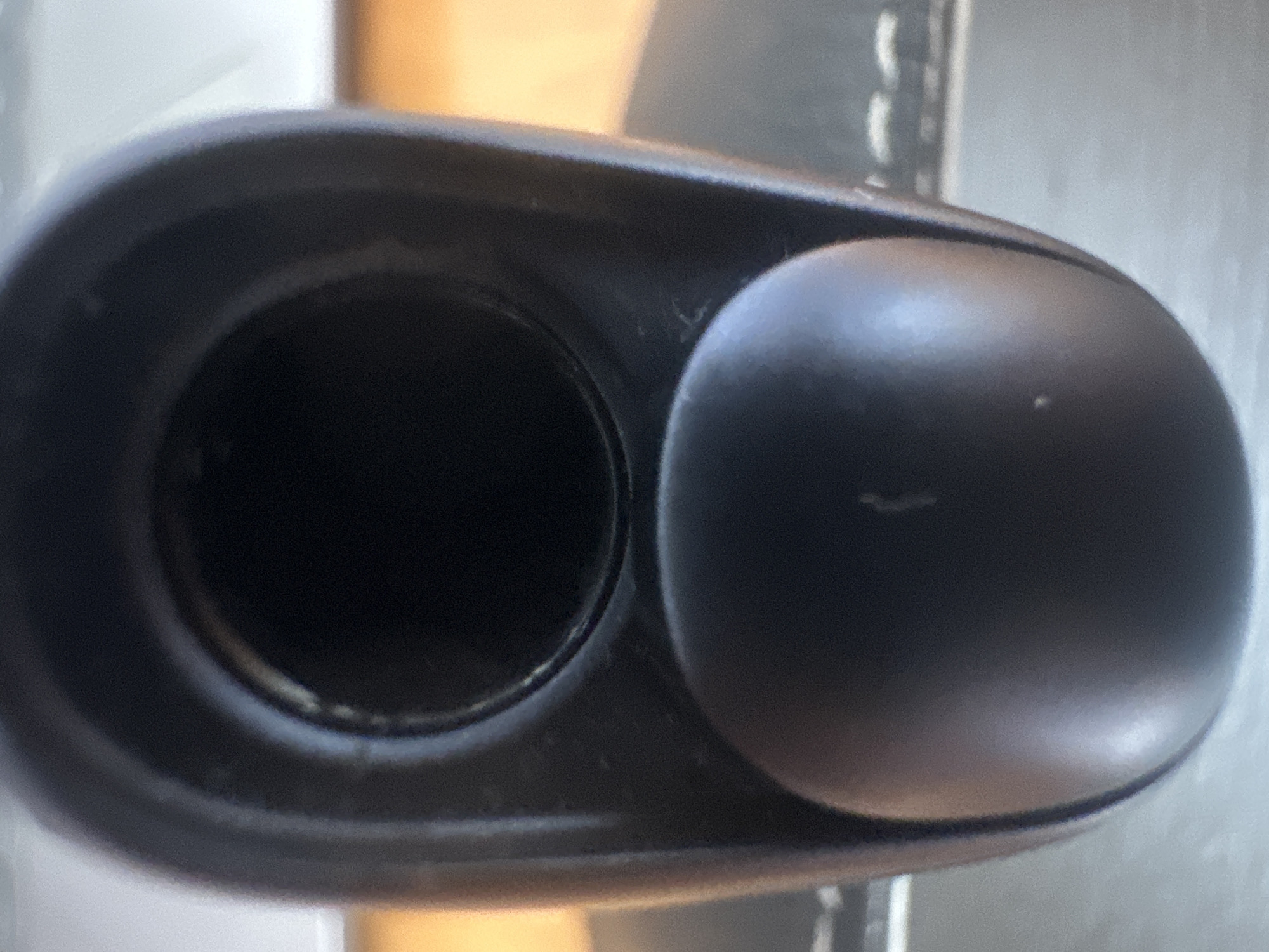 The image shows a close-up, macro view of a black object with two distinct openings. The object has a smooth, matte black finish. The left opening is circular and appears to be a hollow tube or cavity, with some faint lighter-colored residue visible on its inner edge. To the right of this opening is a rounded, bulbous protrusion. The lighting in the image creates soft highlights and shadows, suggesting an indoor environment. The background is out of focus, with areas of muted yellow and gray, providing little contextual information about the setting. There are no people or text visible in the image. The image focuses entirely on the details of the black object, its texture, and its form.