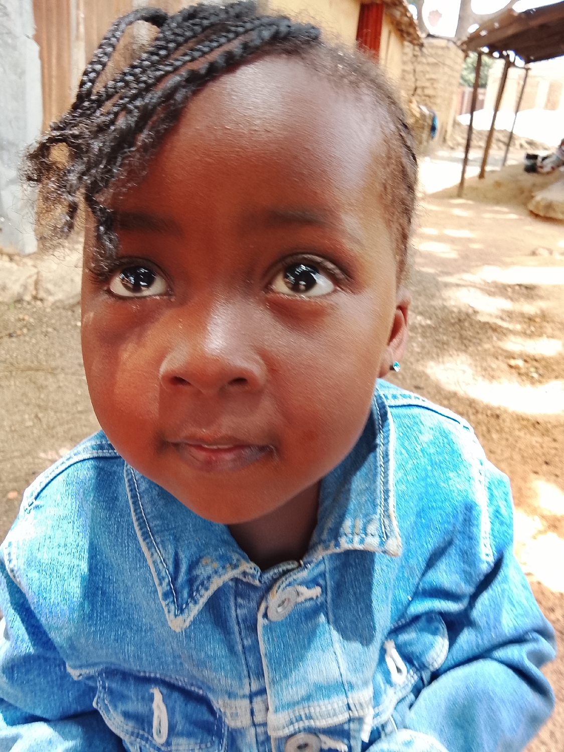 A close-up, eye-level portrait features a young child, likely a girl, with dark skin and prominent dark eyes looking upwards and slightly to the right. Her hair is styled in small braids, with some loose, curly strands framing her forehead. A small, light blue stud earring is visible in her right ear.
The child is wearing a blue denim jacket with light-colored stitching and white buttons, which appears to be a bit large for her. Her expression is neutral to inquisitive, with a slight pout to her lips. Bright sunlight casts defined shadows and highlights on her face and clothing, suggesting it is daytime, possibly midday or early afternoon.
The background shows an outdoor setting with rustic, light-brown structures, possibly made of mud bricks or natural materials. One structure has a visible red door or frame. The ground is sandy or dusty. To the right, a makeshift shelter with a corrugated roof supported by poles can be seen in the distance, indicating a rural or informal settlement environment. No visible text is present in the image.