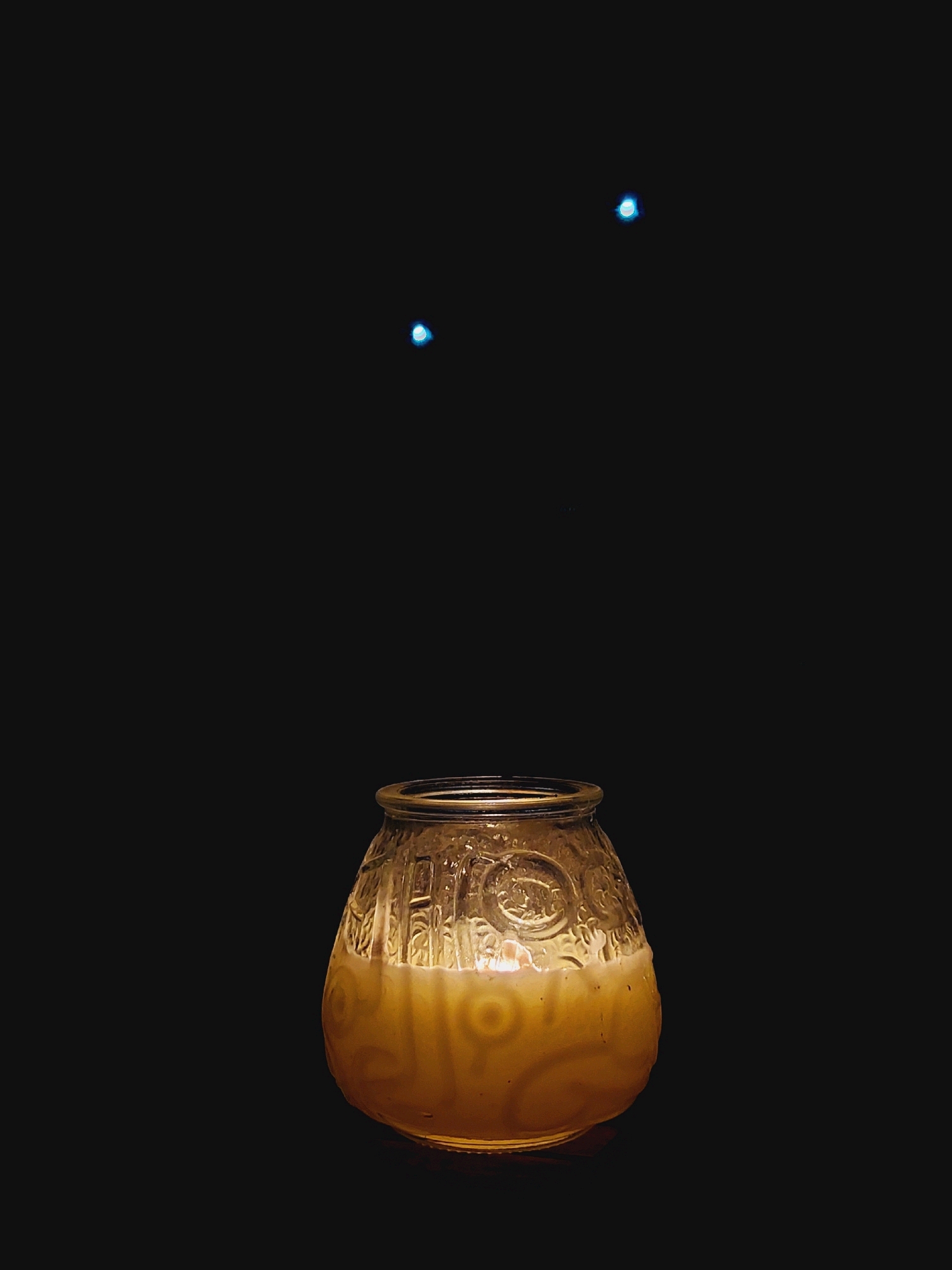 In a dark, possibly outdoor setting, a glass jar filled with a creamy, light-colored liquid sits on a surface. The jar has a textured pattern with circular motifs. Above the jar, three small, bright blue lights are visible, two closer together near the top and one slightly lower and to the left. There is no discernible location in Sozopol, Bulgaria.