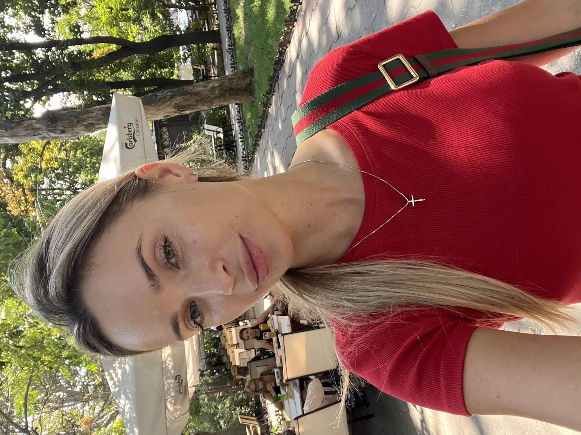 A medium shot captures a blonde woman with green eyes taking a selfie outdoors in Odesa, Ukraine. She is wearing a red ribbed t-shirt and a silver cross necklace. A striped green and red bag strap is visible over her shoulder. In the background, several tables and chairs are set up under a white umbrella, suggesting an outdoor cafe. Two women are seated at a table in the mid-ground, with drinks in front of them. Trees and a paved walkway are also present in the scene. The image is taken in daylight.