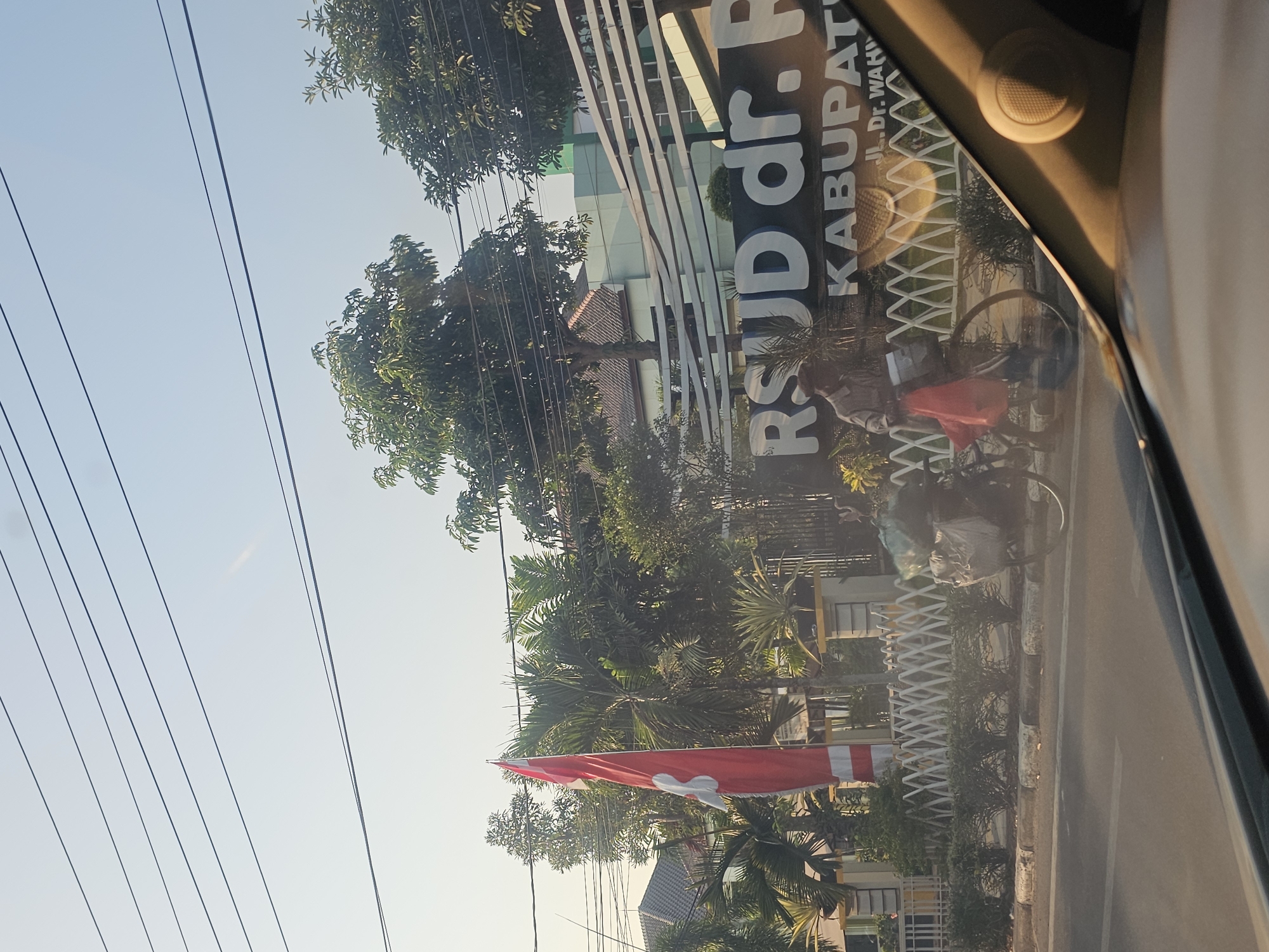 The image shows a street scene in Tuban, Indonesia, in front of RSUD dr. R. Koesma Tuban hospital. A man on a bicycle with a load of items wrapped in plastic bags is visible on the side of the road. Behind the man is a white picket fence and lush green vegetation, including palm trees. A large red and white flag is planted to the left of the fence. The hospital building, with its distinctive white slatted facade, is visible in the background. The sky is clear and a bright sun is casting shadows, suggesting it is daytime. The foreground shows the interior of a vehicle, with a dashboard and speaker visible.