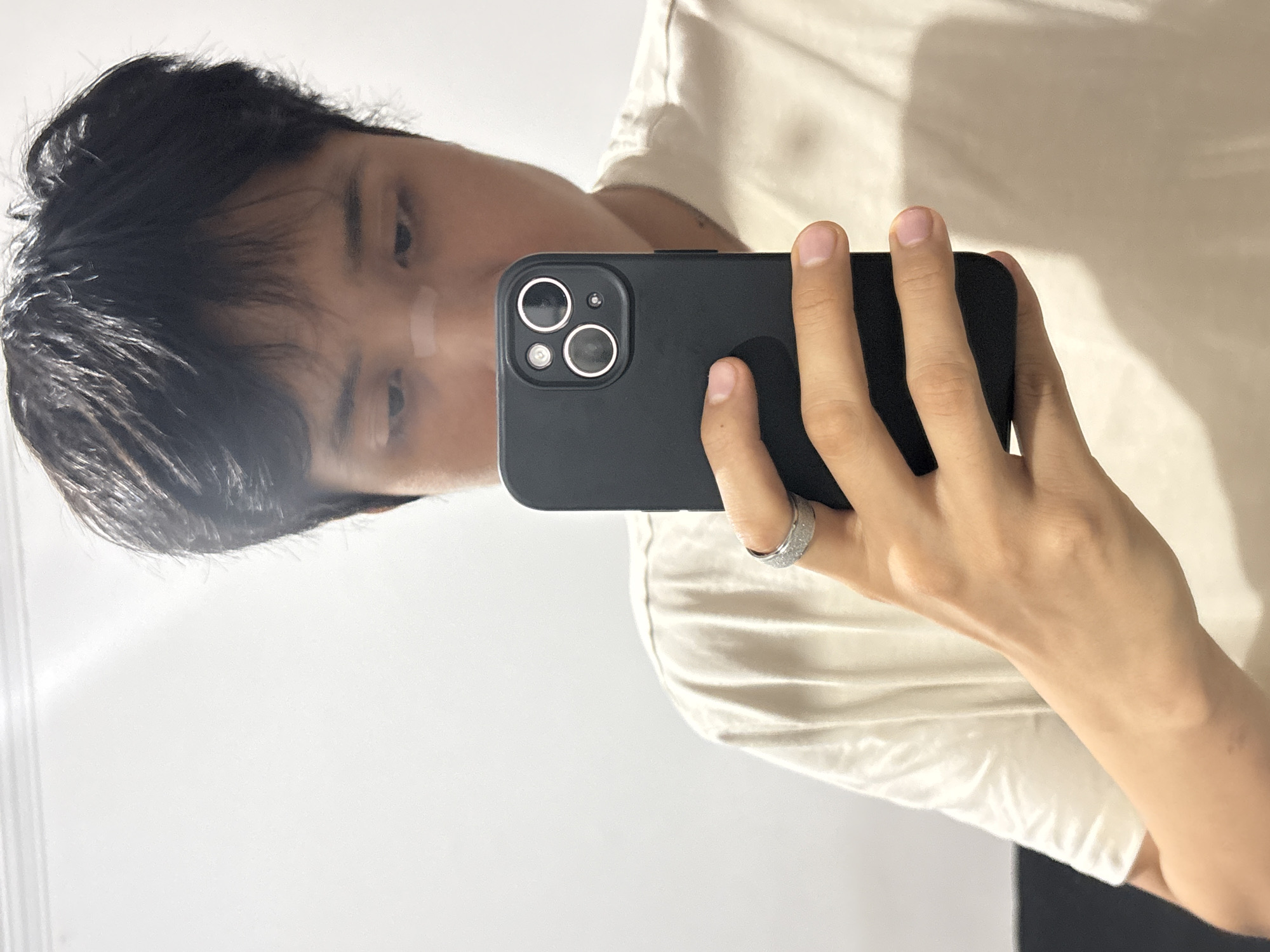 A person with dark hair is holding a black smartphone in front of them. The phone has three camera lenses visible. The person is wearing a white shirt and a ring on their finger. The background is white. The image is taken in a selfie style. There is nothing in the image that indicates the location of the city of São Paulo, Brazil.