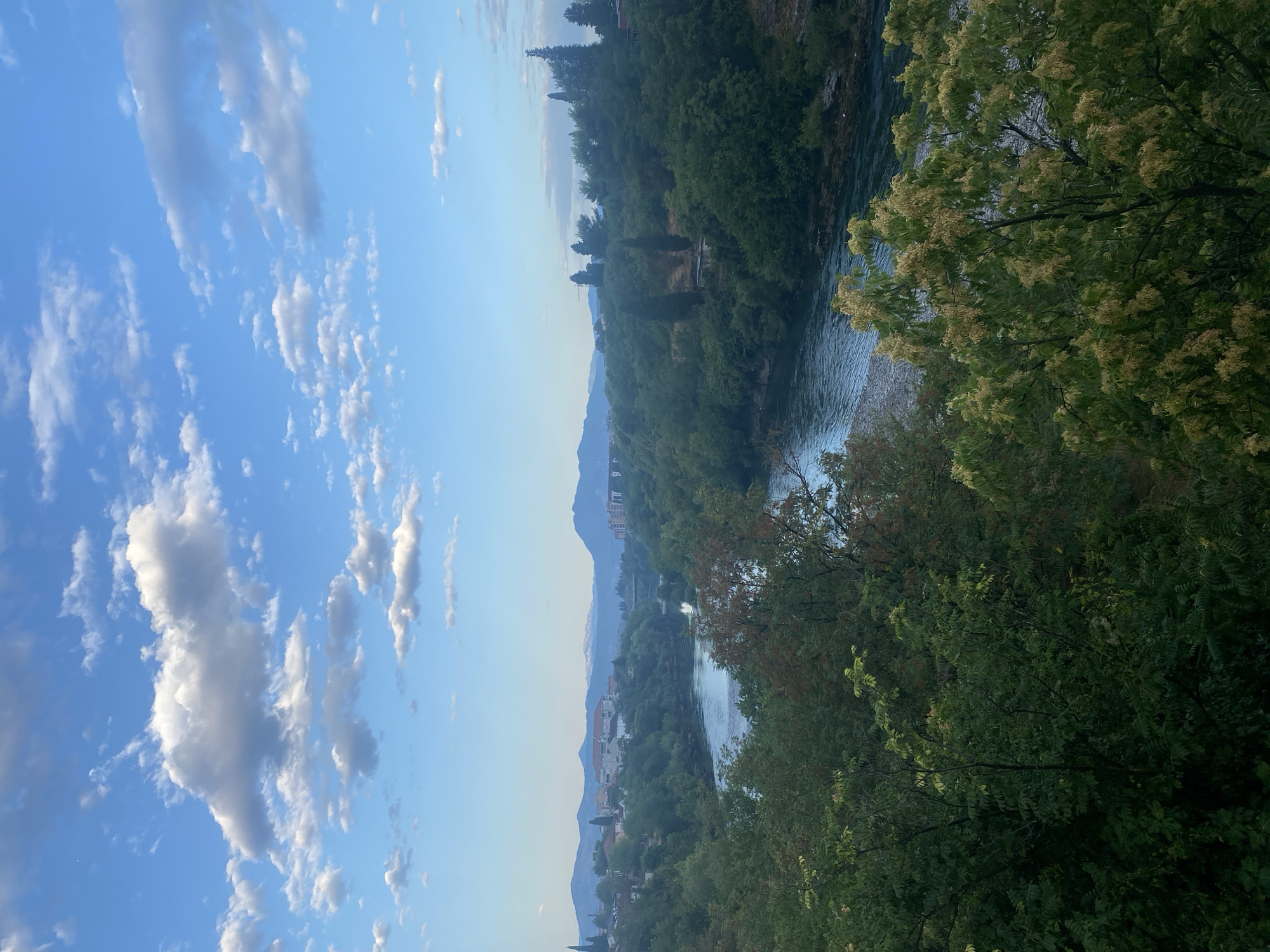This is an eye-level, vertical, full shot of a wide, calm river flowing through a valley filled with dense green trees. The river is a deep blue-gray and has a slightly rippled surface. The trees are lush and varied, with some showing hints of red and yellow foliage, suggesting it might be late summer or early autumn. In the background, on the left side of the river, a town with red-roofed buildings is nestled amongst the trees. Further in the distance, rolling hills and mountains form the horizon, with a haze obscuring their peaks. The sky is a clear, bright blue, dotted with scattered white clouds. This scene is likely the Morača River in Podgorica, Montenegro, possibly viewed from a higher vantage point overlooking the river and the city.
