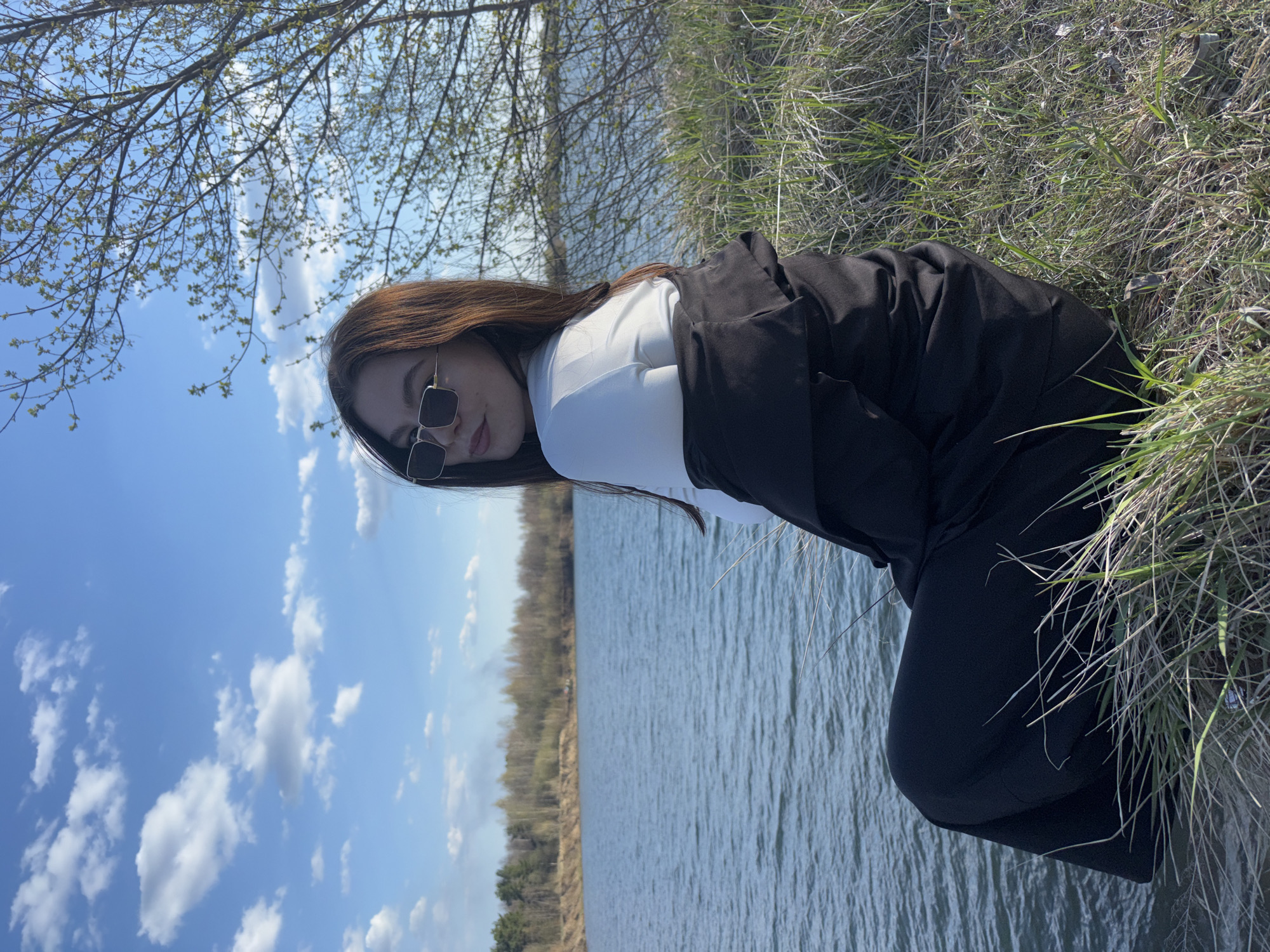 A woman is kneeling near a body of water, possibly a lake, in an outdoor setting. She is wearing sunglasses, a white shirt, and a black jacket with matching pants. The environment consists of grass and foliage, with trees visible in the background. The sky above contains blue patches with scattered clouds. Given the city specified is Zaosinovo, Russia, it is likely that the picture was taken there.