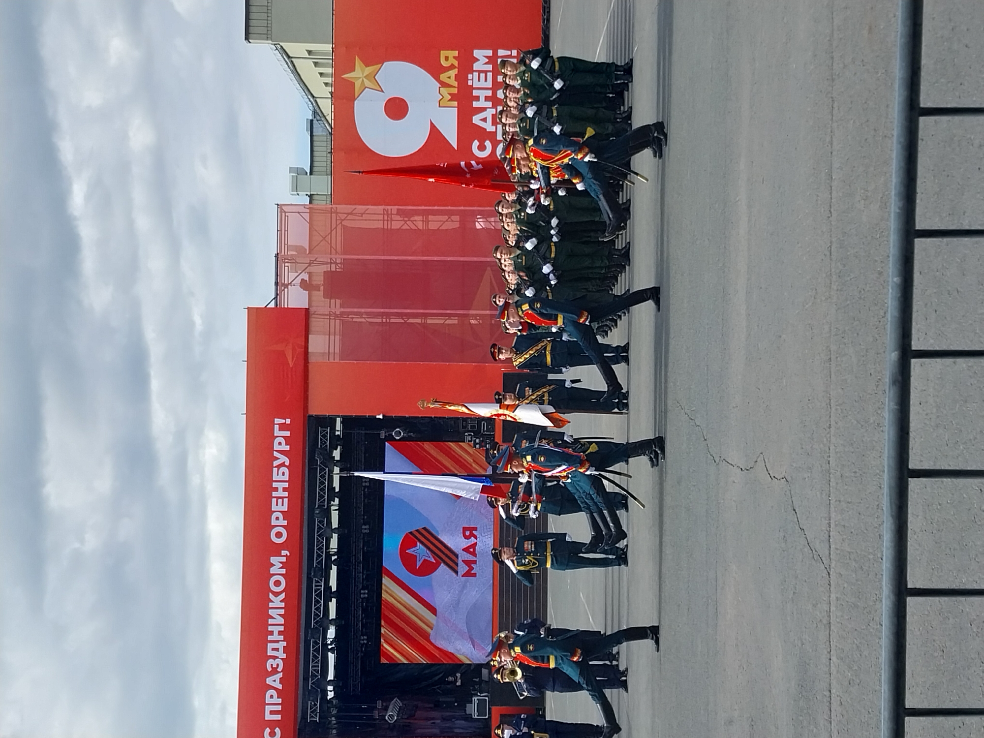 The image depicts a military parade in Orenburg, Russia. Soldiers in dark green uniforms and black boots march in formation on a paved surface, some carrying flags and swords. The background features a large red banner with the words "С ПРАЗДНИКОМ, ОРЕНБУРГ!" (Happy Holiday, Orenburg!) vertically written, and a stage with a screen displaying a stylized "9 МАЯ" (May 9th) design, referencing Victory Day celebrations. The sky above is overcast with clouds. A metal structure is visible on the right edge of the frame.