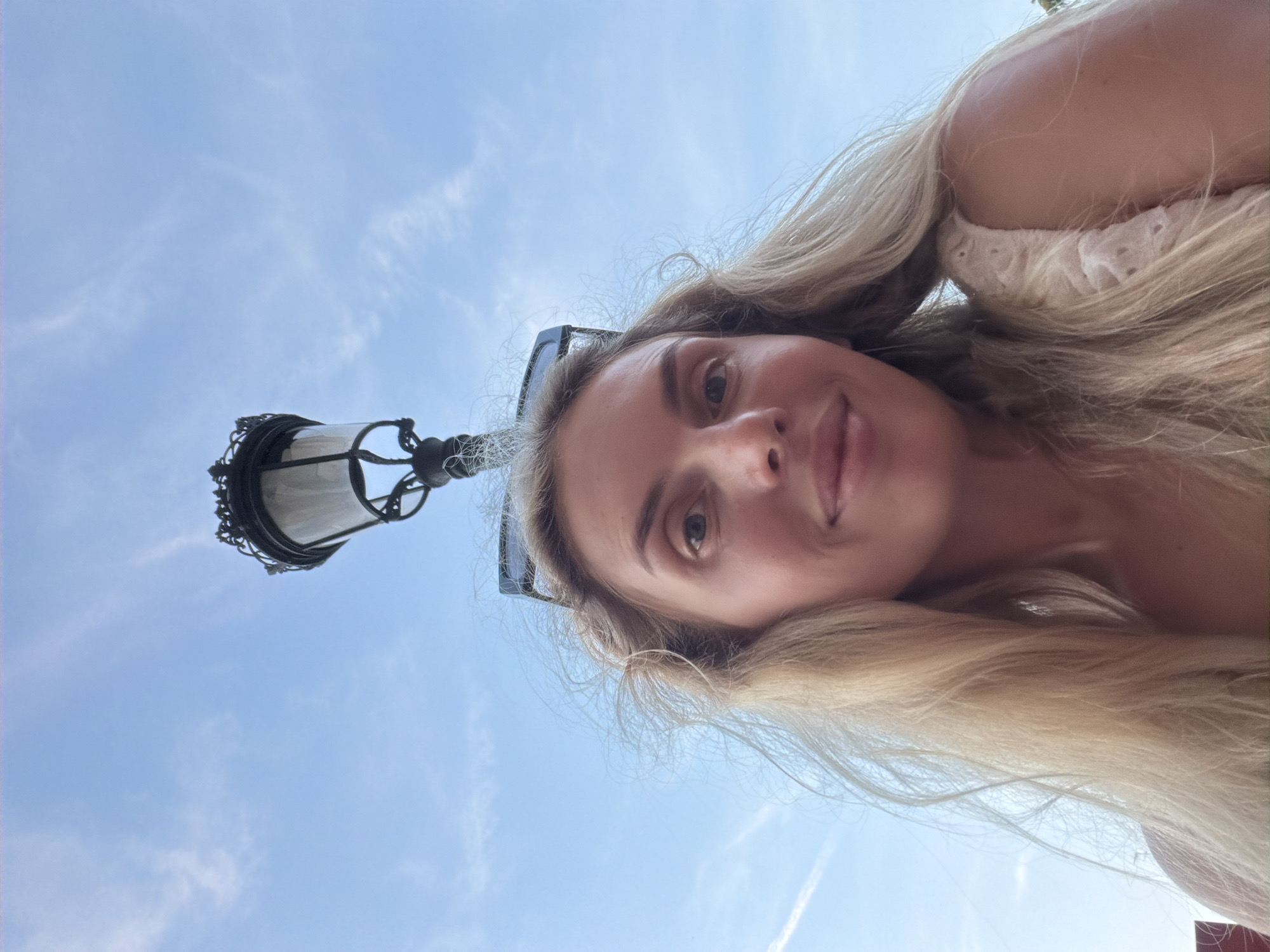 A low-angle, head-and-shoulders selfie of a blonde woman is captured against a bright blue sky with scattered clouds. She is looking directly at the camera with a slight smile, her hair blowing in the wind. A decorative black lamppost is positioned directly above her head, partially obscuring the sky. Sunglasses are perched on top of her head, and she is wearing a light-colored, textured top. The image appears to be taken outdoors, possibly in a public space within Benimàmet-beniferri, Spain, given the presence of a street lamp and clear sky, suggesting a daytime setting.