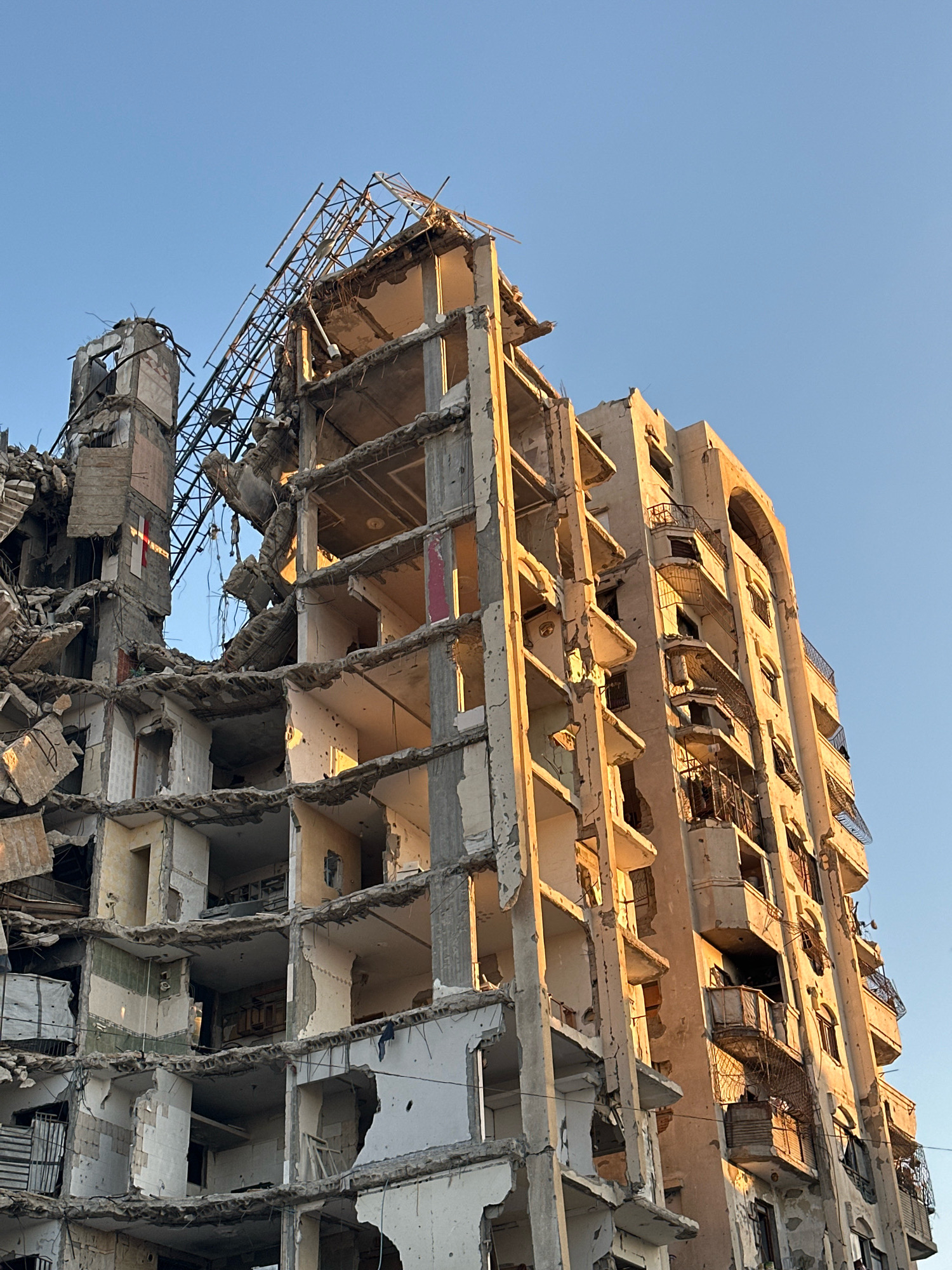 The image depicts the extensive damage to a multi-story apartment building in the Gaza Strip, Palestinian Territories. The building is heavily damaged, with its structure exposed and many sections in ruins. Some floors have completely collapsed, and others show significant structural compromise. Debris is scattered throughout the scene.
The building's facade is marred by damage, with portions of the exterior walls missing, revealing the internal structure of apartments. Balconies are bent and damaged, some with remnants of railings. The windows are mostly gone, leaving open frames.
In the background, a clear blue sky is visible. The lighting suggests it is either early morning or late afternoon, casting warm tones on the damaged structure. There are no people visible in the image. The overall scene is one of destruction and devastation, characteristic of conflict.
