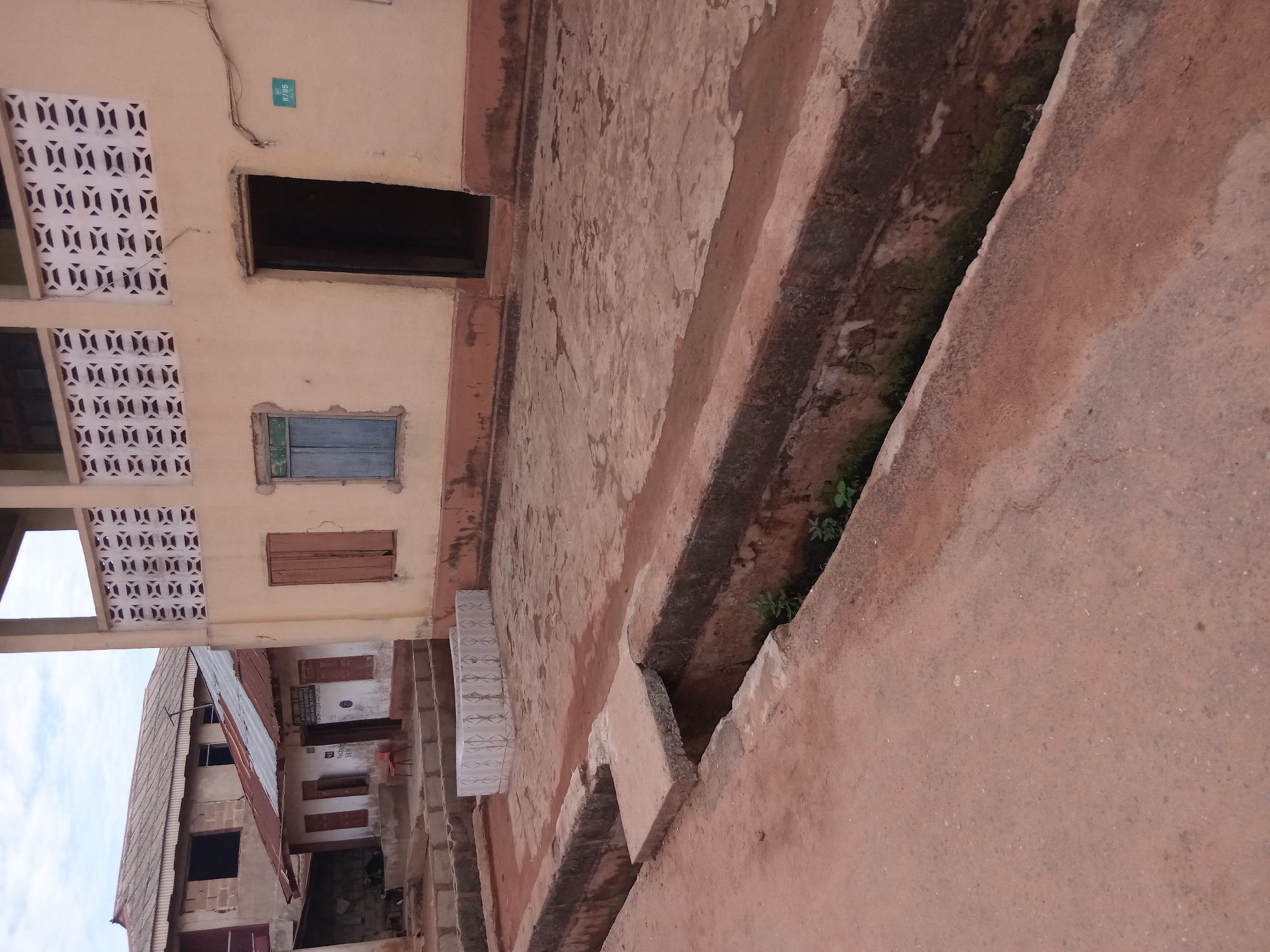 The image shows a street scene in Shagamu, Nigeria. A two-story building with decorative concrete latticework is visible on the left. The building has windows, one with a blue shutter and another with a brown shutter. There is a large opening in the wall where a window may have been. The building is labeled "K/35". Adjacent to this building, another building with a corrugated metal roof is visible. An elevated walkway or platform, possibly made of concrete, runs along the right side of the image. A drainage ditch filled with some vegetation is embedded in the walkway. The ground appears to be reddish brown. There are no people visible in the image.