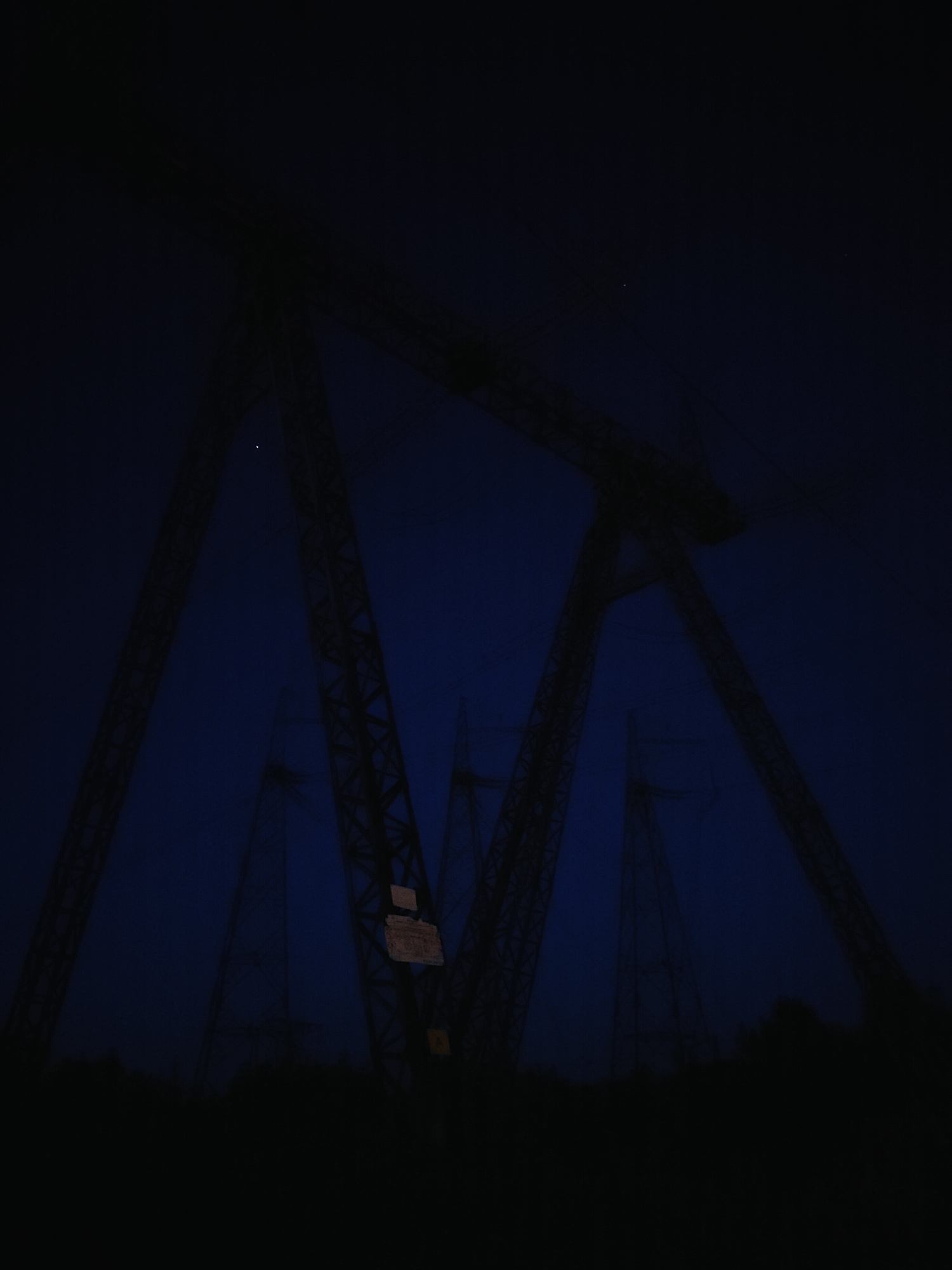 The image depicts a low-angle view of tall, dark electricity pylons against a deep blue, twilight sky. The pylons are lattice steel structures, appearing silhouetted and somewhat blurred due to the low light conditions. Some wires are visible stretching between the pylons. In the distance, other similar pylons are faintly visible, also against the dark sky. The ground at the bottom of the image is obscured by darkness, suggesting it is nighttime or dusk. There is a sign attached to one of the pylons, but its text is illegible. The scene appears to be an industrial area or a landscape with significant power infrastructure. While the exact location within Chaikovsky, Russia, is not explicitly identified, the presence of these structures is characteristic of such regions.