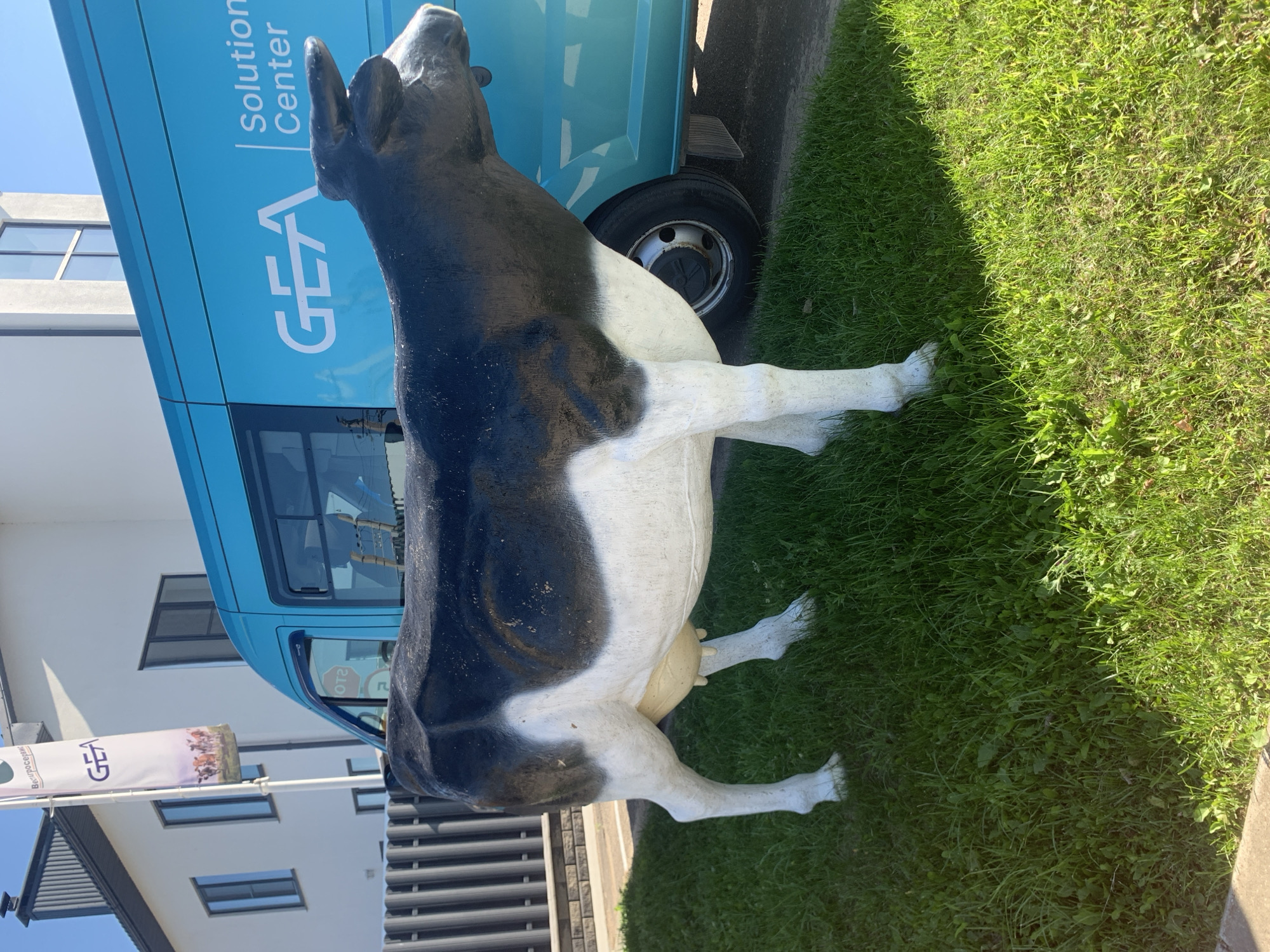 A life-sized black and white cow statue stands on a bright green lawn in front of a turquoise van. The van has "GEA Solution Center" written in white on its side, along with a GEA logo. Behind the van, a white building with windows and a dark roof is visible, and a tall white flag with the GEA logo and images of cows is planted on the left. The scene appears to be outdoors in a sunny environment.