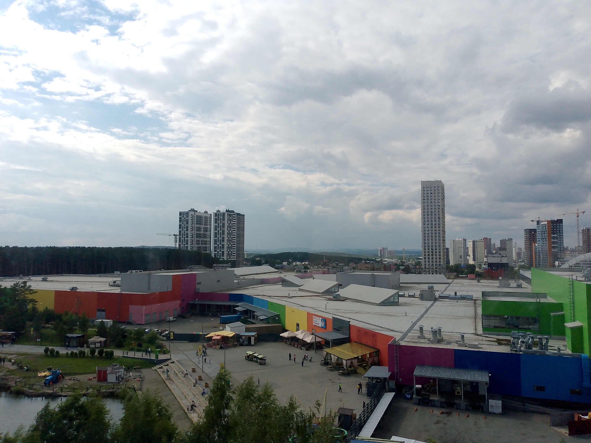 This is an aerial view of a sprawling, modern shopping mall complex with a diverse range of colorful facades. The mall is situated amidst a cityscape with multiple high-rise buildings, including a very tall, slender skyscraper in the background, and a dense forest bordering one side. In the foreground, there is a body of water, likely a lake or river, with a small park area and some children's play structures, including a blue toy tractor. A few people are visible scattered around the park and on walkways leading to and from the mall.
The mall itself is a striking architectural feature with sections painted in bright, bold colors such as yellow, orange, red, pink, green, blue, and purple. Large, flat roofs dominate the structure, punctuated by numerous ventilation systems and skylights. Several smaller structures, possibly kiosks or food stalls, are set up in open areas around the mall, with awnings providing shade. One awning displays a red and yellow logo.
In the distance, the skyline is a mix of modern apartment buildings and older, more industrial-looking structures. The sky is overcast with a mix of white and gray clouds, suggesting a cloudy day. The overall impression is one of a contemporary urban environment with a focus on commercial activity and leisure, possibly in a suburban or outskirt area of a city. The scene appears to be set during daylight hours, likely late morning or afternoon, given the lighting conditions. There is no discernable text in the image.