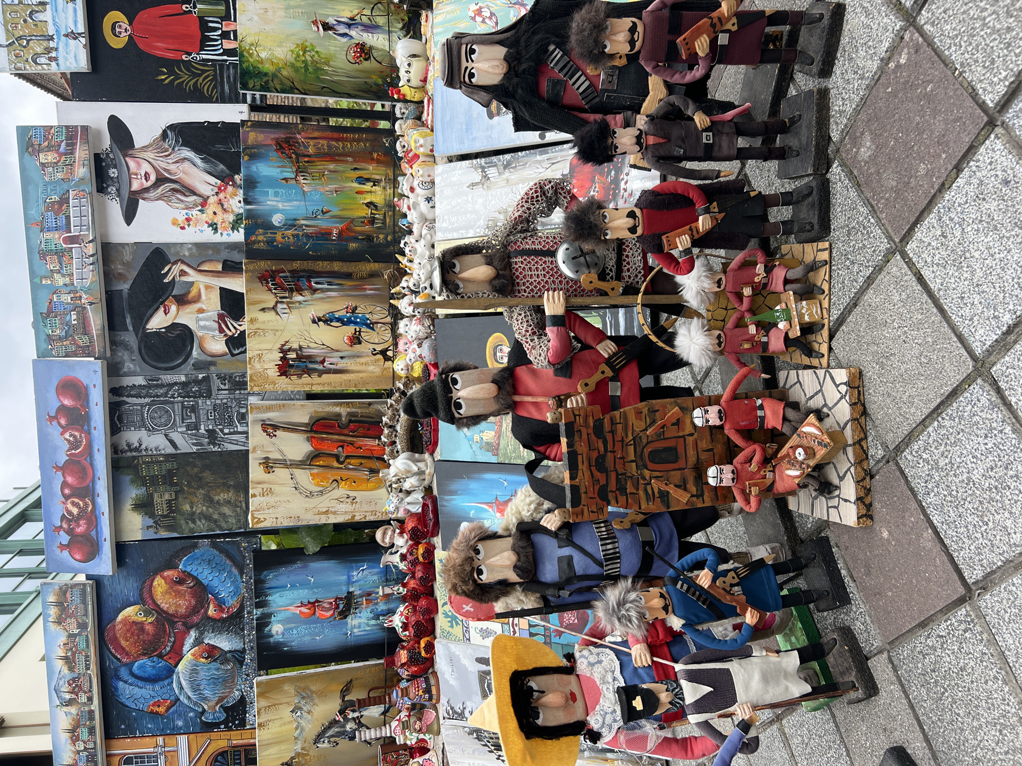 This image depicts a street market scene in Tbilisi, Georgia, likely in the Old Town area, showcasing a collection of handcrafted dolls and paintings. The dolls are diverse, featuring male and female figures dressed in traditional or stylized clothing. Many of them have distinct facial features, such as prominent noses and moustaches, and some wear hats or head coverings. The dolls are arranged in groups and individually, with some holding props like rifles, swords, or musical instruments. There are also miniature structures, such as a brick tower and a small table with figures seated around it.
Behind the dolls, a variety of paintings are displayed, creating a colorful backdrop. These paintings exhibit different subjects, including landscapes of cities, still lifes with pomegranates and fish, portraits of women, and depictions of musical instruments. The overall atmosphere suggests a vibrant marketplace where local crafts are being sold. The ground is paved with stone tiles, common in pedestrian areas of historic cities.