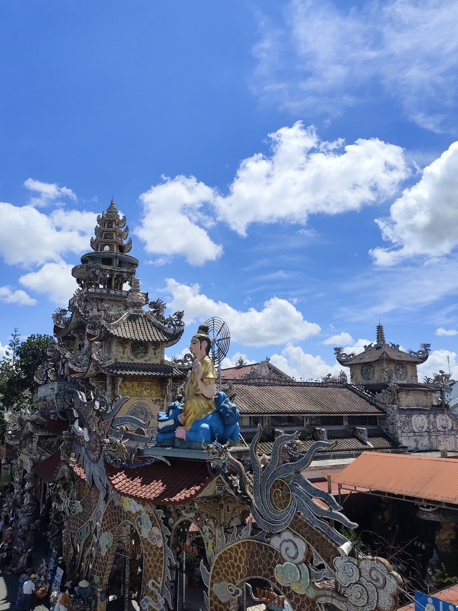 The image captures a detailed view of a Buddhist temple, likely the Linh Phuoc Pagoda, located in the city of Xuân Thành, Vietnam. The architecture is intricate and ornate, featuring a multi-tiered pagoda structure adorned with what appear to be ceramic mosaic decorations. Dragon sculptures are prominently featured on the rooflines and surrounding the structure, a common motif in Vietnamese temples.
In the foreground, a statue of a person in golden robes sits atop a blue creature, possibly a dragon or a mythical beast. Behind this, the main pagoda rises, with a smaller, similar structure visible further in the background, also capped with a tiered roof. The roofs are tiled in a traditional style. The sky above is a vibrant blue, punctuated by fluffy white clouds, suggesting a clear day.
The lower section of the image shows a glimpse of the temple grounds, with several people in casual attire walking and interacting. Some are carrying baskets, possibly for offerings or shopping. The overall impression is one of a richly decorated religious site, bustling with activity under a bright sky.