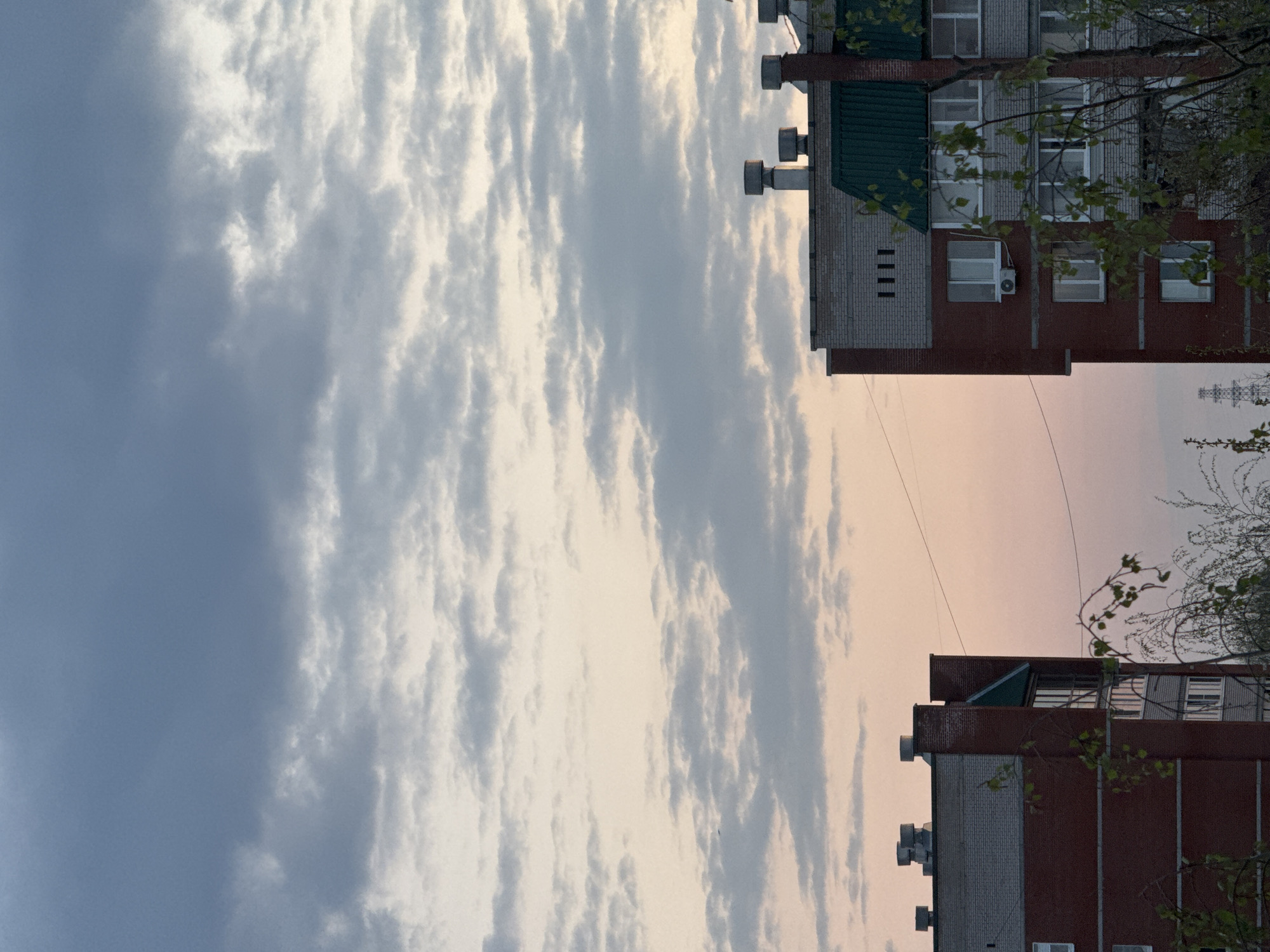 The image shows two multi-story buildings in Chaikovsky, Russia, under a sky filled with striated clouds. The buildings are brick and white brick, with visible windows and air conditioning units. Bare tree branches are visible on the sides of the buildings. A power line stretches between the two buildings. The sky varies from dark blue to a pale orange-yellow.