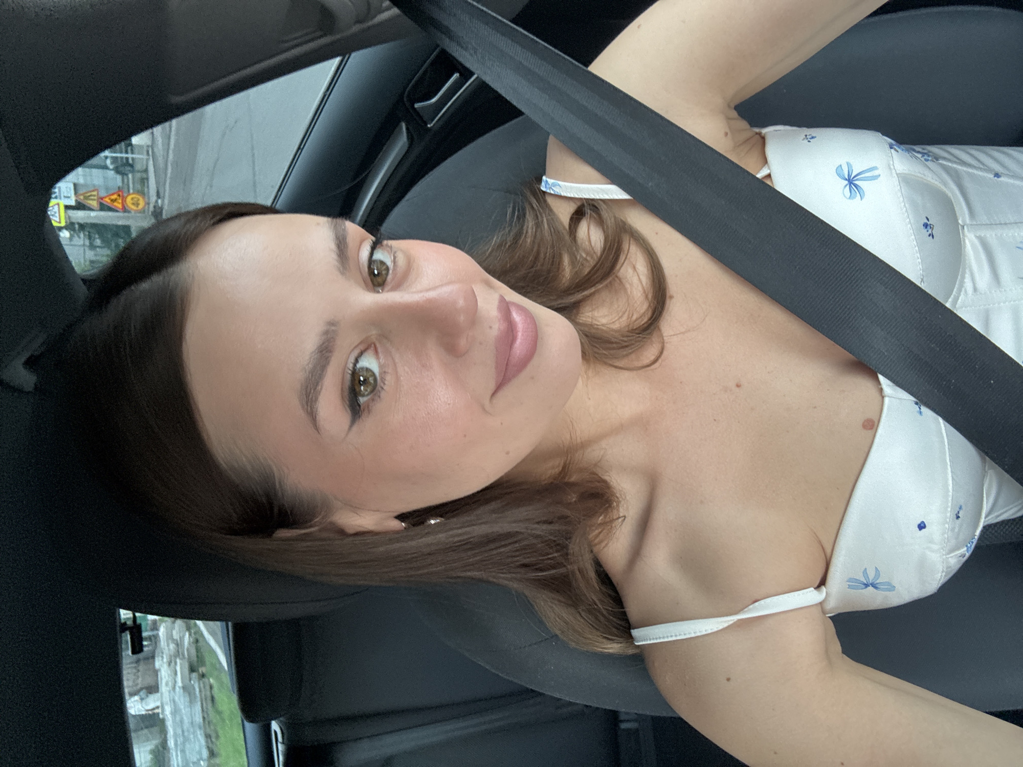 A young woman with light brown hair and green eyes is sitting in the driver's seat of a car, smiling at the camera. She is wearing a white, satin camisole with a blue floral pattern. A black seatbelt is fastened across her chest. Outside the car window, a street is visible with trees and buildings, as well as traffic signs including a speed limit sign indicating 40. The background suggests an urban environment.