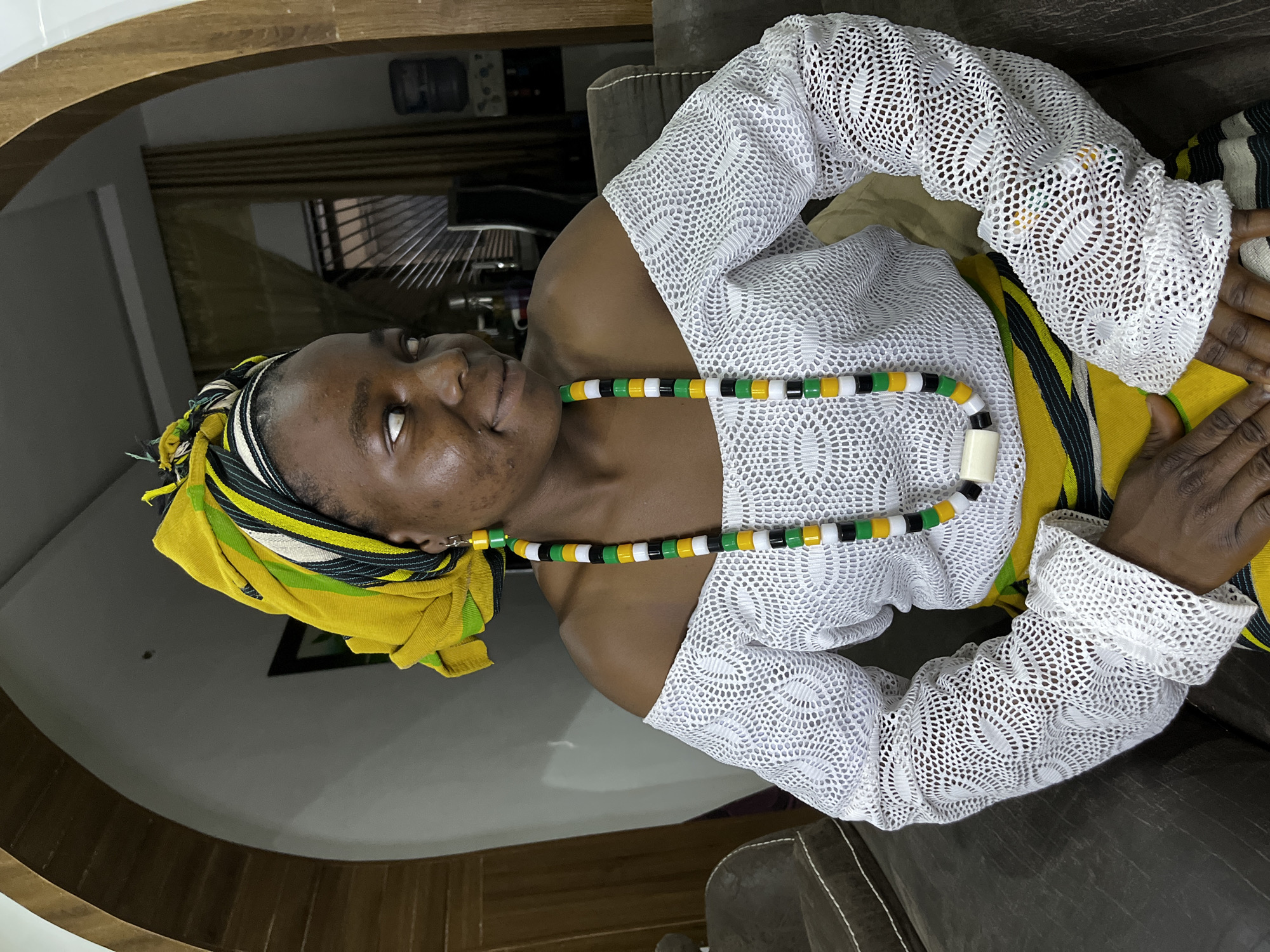 A medium shot captures an individual with brown skin in an interior setting. The person is wearing a yellow headscarf patterned with green, black, and white stripes. They also wear a white, long-sleeved top made of lace fabric, along with a beaded necklace of green, yellow, white, and black. The subject's hands are visible, resting on a lap covered by striped fabric. The person is situated on a dark-colored upholstered chair. The location is Jos, Nigeria. The environment appears to be a residential interior with muted lighting.
