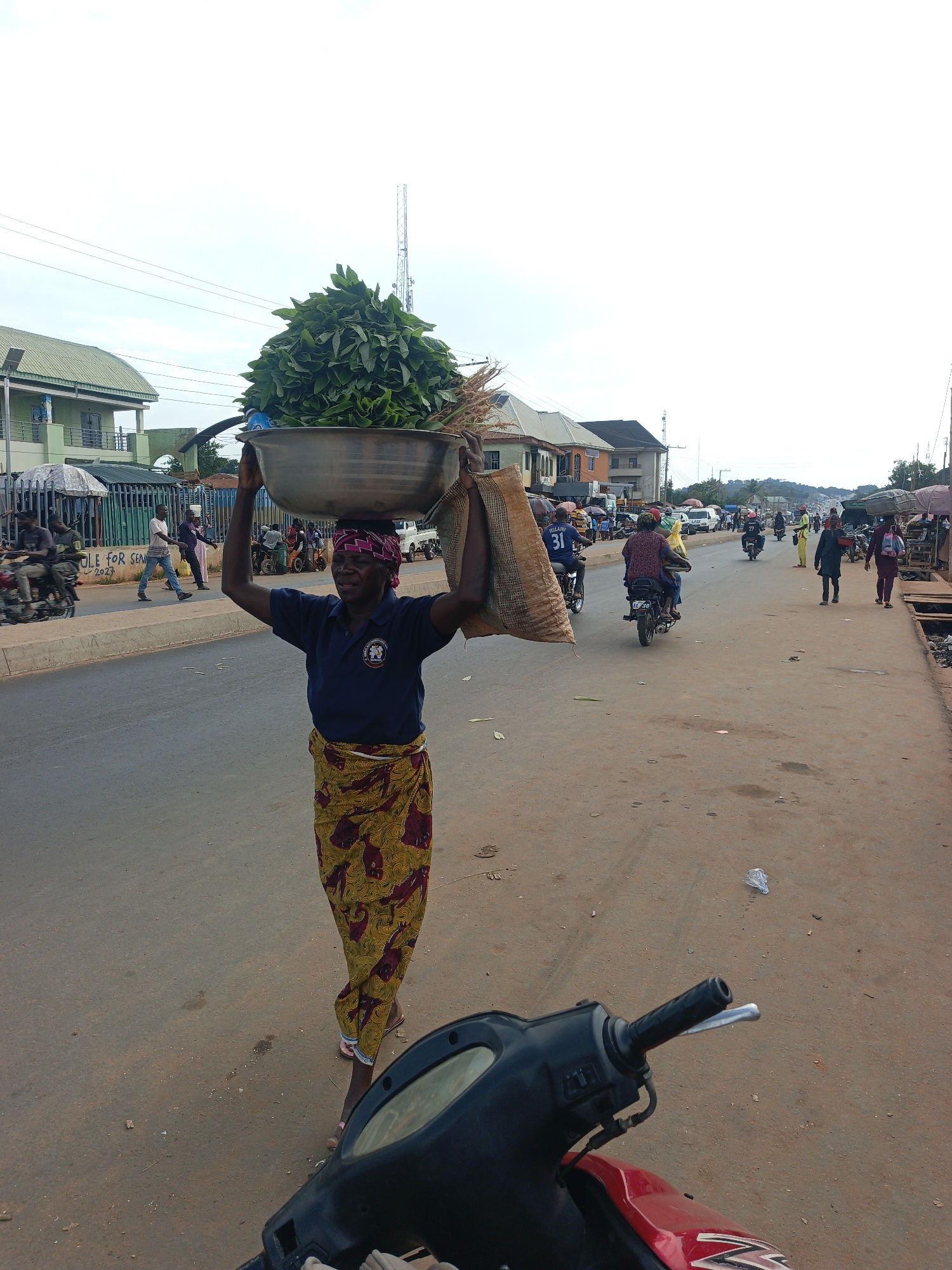 An eye-level, outdoor, daytime shot shows a woman carrying a large metal bowl filled with leafy green produce and a bag of goods on her head as she stands on a busy street in Akwanga, Nigeria. The woman is dressed in a dark blue, short-sleeved shirt, a patterned yellow and red wrap skirt, and wears a red patterned headscarf. She is centered in the frame. The foreground includes part of a red and black motorcycle, slightly out of focus. The middle ground shows a paved street with other people walking and riding motorcycles. In the background are buildings, utility poles, and a cloudy sky. Street vendors can be seen on both sides of the street.
