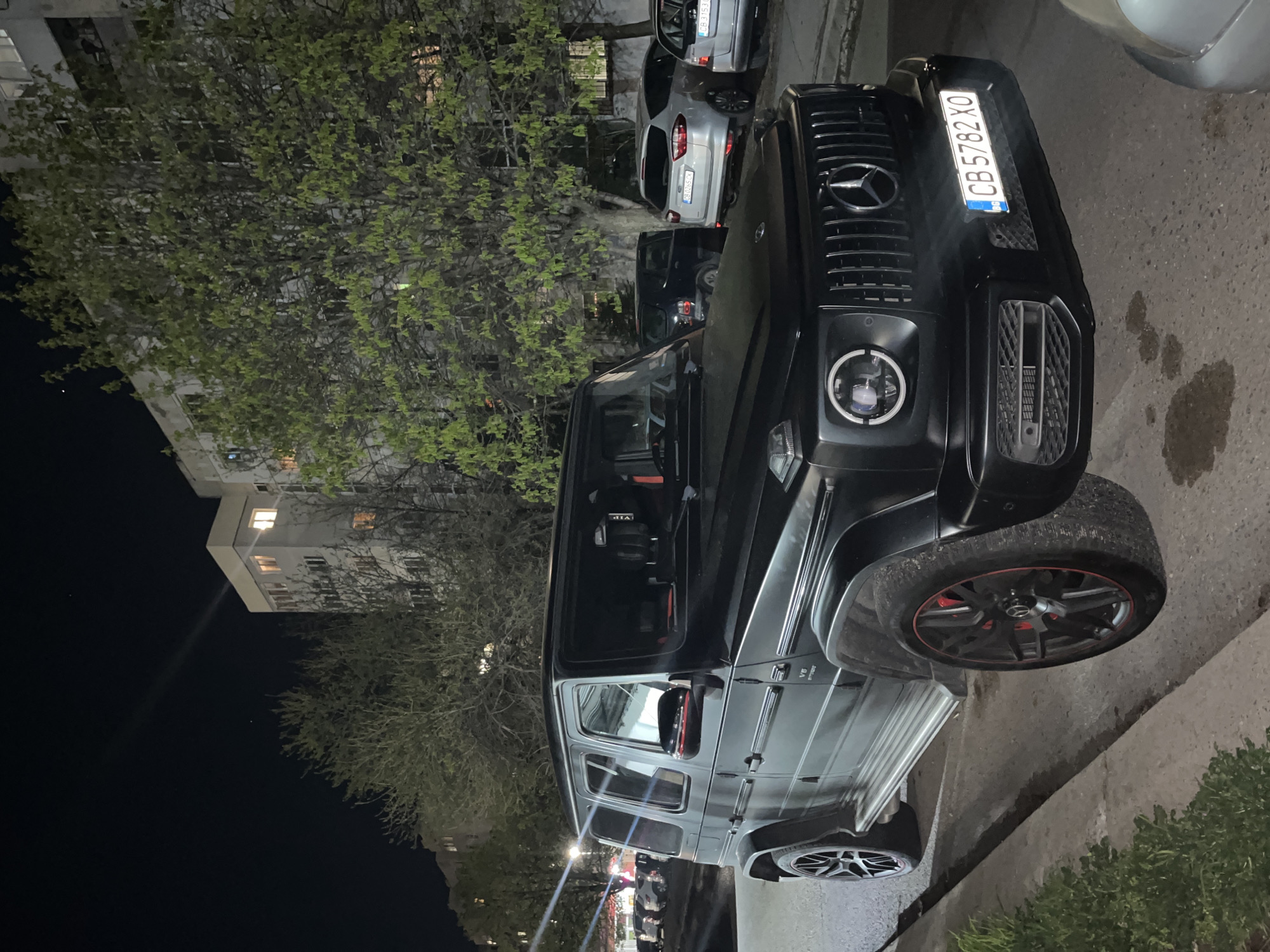 The image shows a black Mercedes-Benz G-Class, specifically a G63 AMG, parked on a street in Sofia, Bulgaria. The license plate number is CB 5782 XO. The car is parked near a sidewalk and a tree. In the background, there is a building with lit windows, indicating it's nighttime. Other cars are visible parked behind the G-Class. The focus is clearly on the car, highlighting its front design, including the headlight and grille.