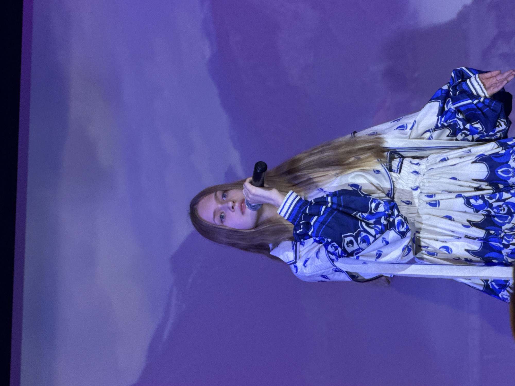 A woman with long, light brown hair is holding a microphone and looking directly at the viewer. She is wearing a white dress with a blue floral pattern and white and blue striped cuffs. The background is a soft purple color. The location is Unknown, Unknown.