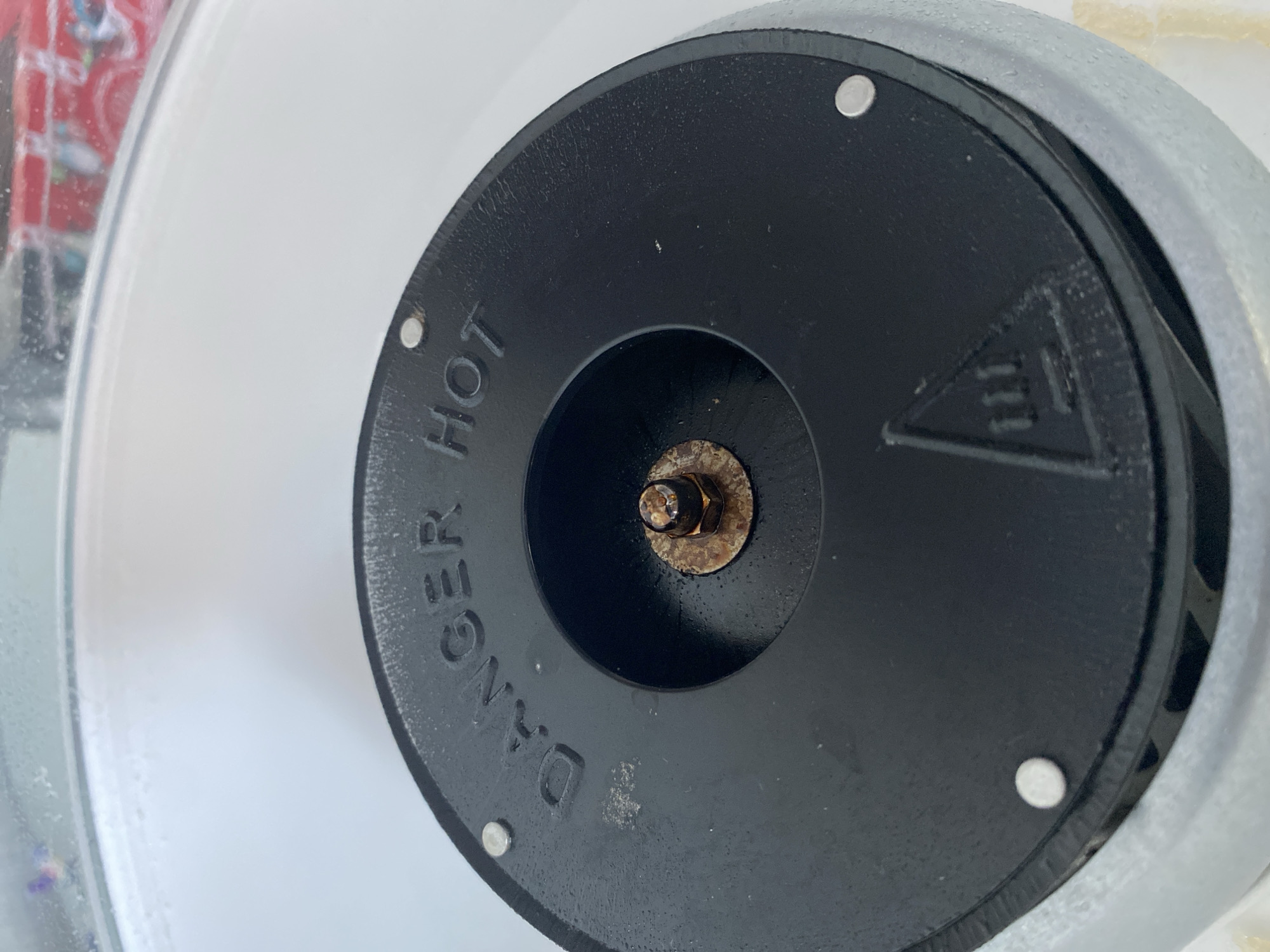 The image shows a close-up, overhead view of a black, circular object with embossed text that reads "DANGER HOT." The object has several small, silver-colored fasteners around its perimeter. In the center of the black object is a recessed area with a rusted bolt and nut. A triangular symbol with three vertical lines inside it is visible on the lower right portion of the black surface. The object appears to be part of a larger, white appliance, possibly a cotton candy machine, given the "DANGER HOT" warning and the central spinning mechanism implied by the bolt. The surrounding environment is out of focus but appears to be a bright, indoor setting with some water droplets visible on the upper edge of the appliance, suggesting it may be humid or has recently been cleaned. There are no people or animals in the image. The location in the city of Balashikha, Russia, is not determinable from the visual information provided in the image.