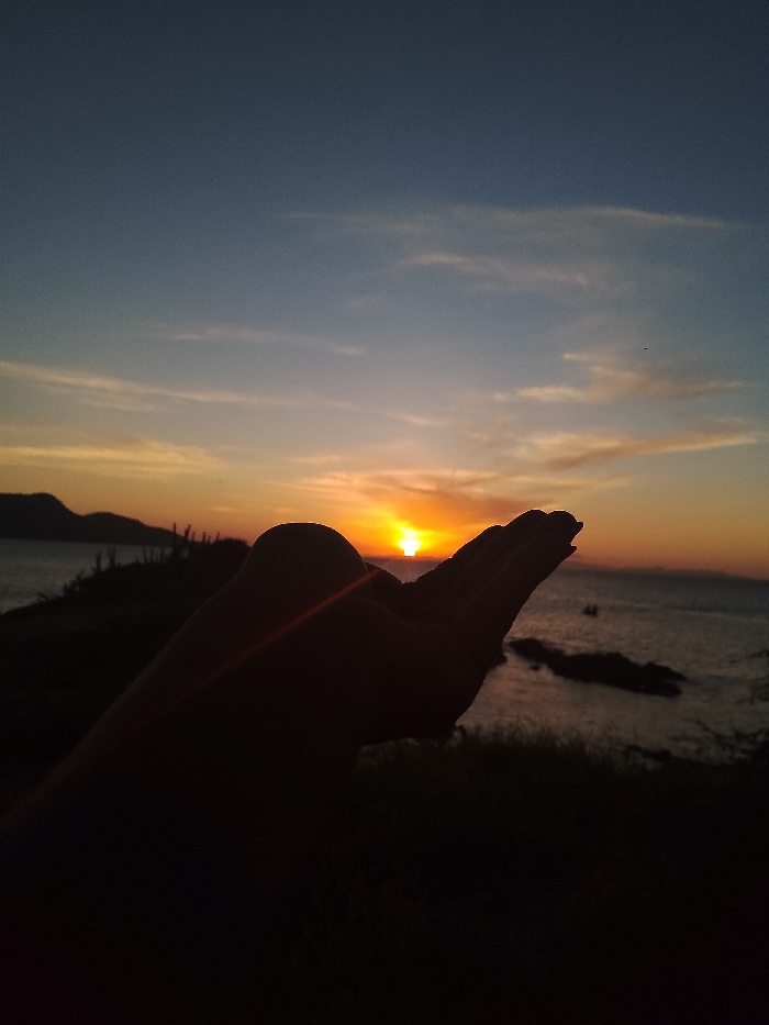 A striking silhouette of a hand appears in the foreground, its palm open to cradle the setting sun. The sun, a bright orb, rests perfectly within the hand's reach, casting a warm glow. The scene unfolds over a calm ocean, dotted with rocky formations. Distant landmasses frame the horizon, silhouetted against the sunset's hues. The sky transitions from a deep blue to vibrant orange and yellow where the sun dips.