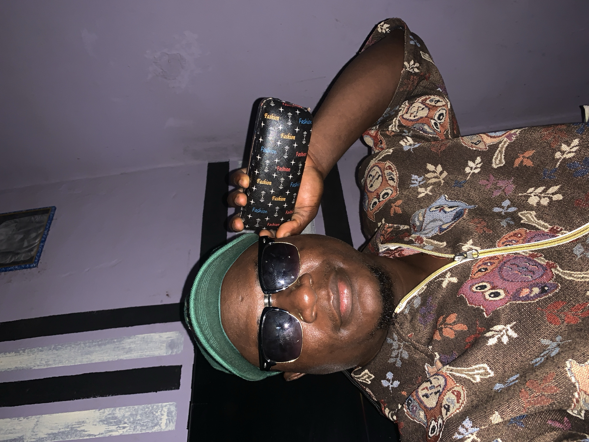 The image features an indoor portrait of an individual, likely male, from the chest up, in Akwanga, Nigeria. The person has dark skin, a short beard, and is wearing a green visor cap backward with dark sunglasses perched on his head. He is dressed in a brown, patterned top or jacket, featuring stylized owls, leaves, and abstract shapes in shades of blue, red, purple, orange, and white. A yellow zipper is visible on the front of his garment.
In his right hand, he holds a black case, possibly for glasses, decorated with white cross patterns and the word "Fashion" repeated multiple times in varying colors such as red, blue, white, and yellow. Some instances of the text appear as "Fashio" or "Fasivon."
The background consists of a light purple or lavender-colored wall and ceiling. The ceiling shows areas of discoloration or texture, potentially indicating marks or uneven plaster. The lower part of the wall is painted with distinct black and white horizontal stripes. A framed object is partially visible on the far left. The lighting is dim, suggesting an indoor setting under artificial light or during evening hours. The person maintains a neutral expression, with his eyes hidden by the sunglasses.