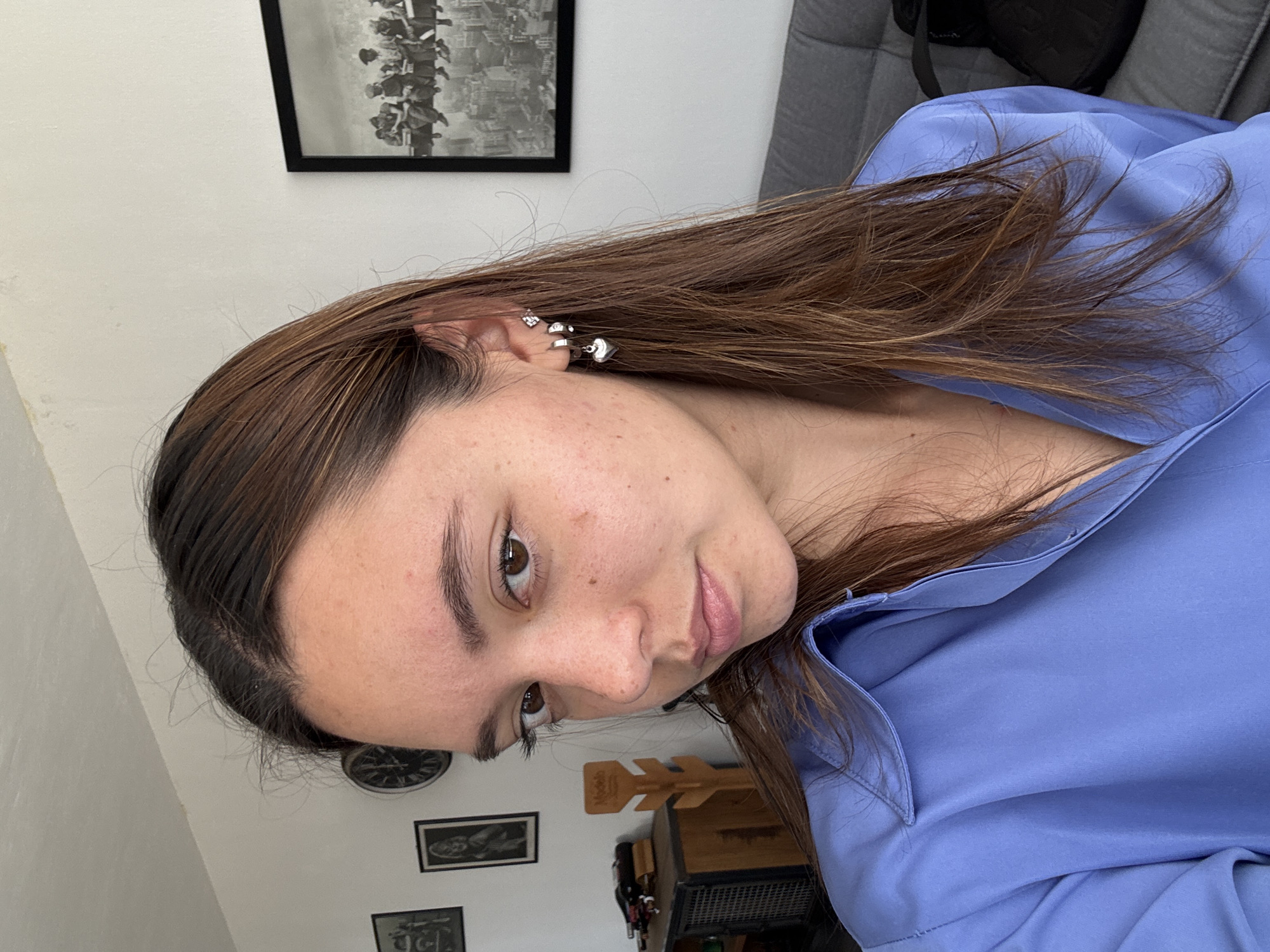 A young woman with medium-length brown hair is posing for a selfie. She is wearing a light blue collared shirt. Her skin has some freckles and minor blemishes. She has several silver earrings in her left ear. Behind her are a gray couch, framed pictures on a white wall, and other miscellaneous objects. The location is not evident from the image, and it's not possible to verify that this photograph was taken in San Bernardino Tlaxcalancingo, Mexico.
