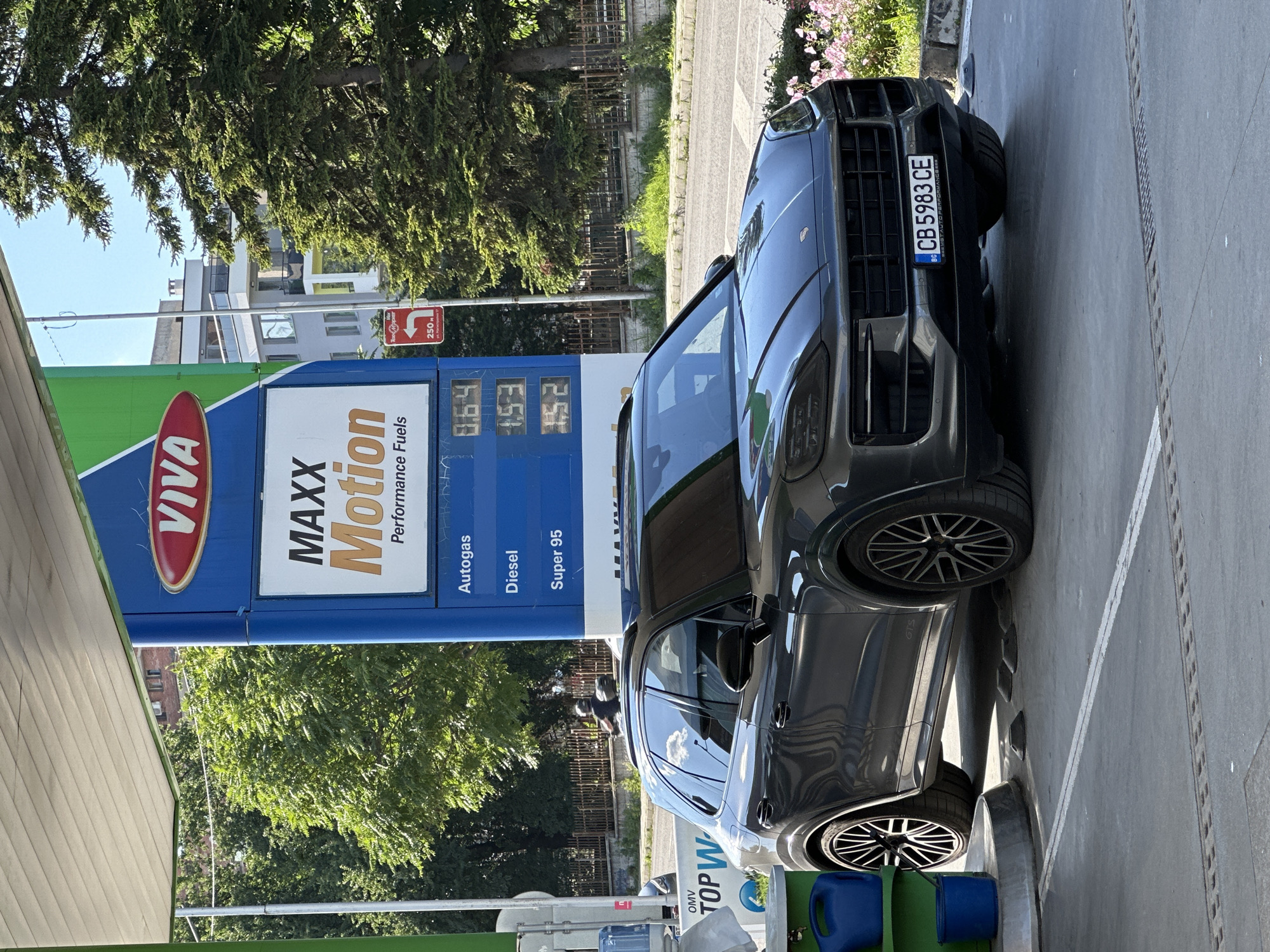 This media file shows a dark gray Porsche Cayenne parked at a VIVA petrol station in Veliko Tarnovo, Bulgaria. The car is positioned diagonally in front of the fuel pumps. The petrol station sign displays "VIVA MAXX Motion Performance Fuels" and prices for Autogas, Diesel, and Super 95 fuel, which appear to be 1.53, 1.52, and 0.64 respectively. The license plate of the Porsche is visible as CB 5983 CE, with "BG" indicating Bulgaria. The sky is clear and blue, suggesting a sunny day. Lush green trees are visible in the background, along with a multi-story building. A person can be seen in the reflection on the car window, and a red directional sign indicating "250 m" and a right turn is also present. Two blue canisters are placed on the ground near the car. The overall impression is a routine stop at a gas station on a bright, clear day.