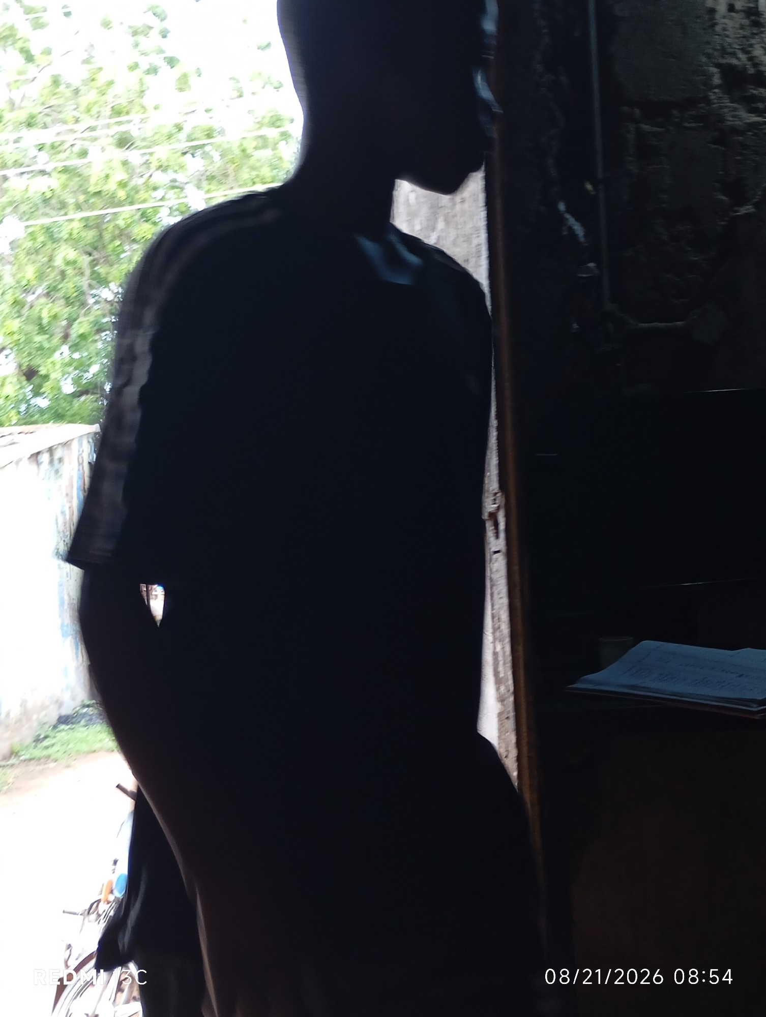 A silhouette of a person is visible in the foreground of the image. The person is wearing a dark-colored shirt. The background shows a mix of indoor and outdoor elements. To the left, there's an outdoor scene with greenery and a wall, suggesting a location in Shendam, Nigeria. To the right, the scene is darker and appears to be indoors, with a wall and a table with a book or papers on it. A timestamp at the bottom right indicates the date and time as 08/21/2026 08:54. There is a partial view of a bicycle in the lower left corner. The image has a low resolution and is grainy.