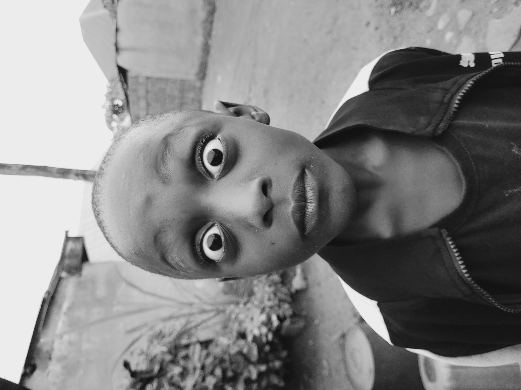 A black and white, medium close-up photograph depicts a young Black child with wide, startled eyes and a slightly open mouth, looking directly at the camera. The child has a shaved head and is wearing a dark jacket with white accents on the shoulders and sleeves, zipped partially open. The background suggests an outdoor, possibly rural or semi-urban setting. To the left, blurred green foliage is visible, along with the top of a cylindrical object. Behind the child, a textured wall or building facade with a dark horizontal band runs across the frame. Further in the background, there are glimpses of rooftops and what appear to be low-lying structures, consistent with the architecture often found in Bukuru, Nigeria. The ground in front of the child is a rough, indeterminate surface. The overall lighting is soft and diffused, suggesting an overcast sky or late afternoon sun. The image appears to be taken at eye level with the child.