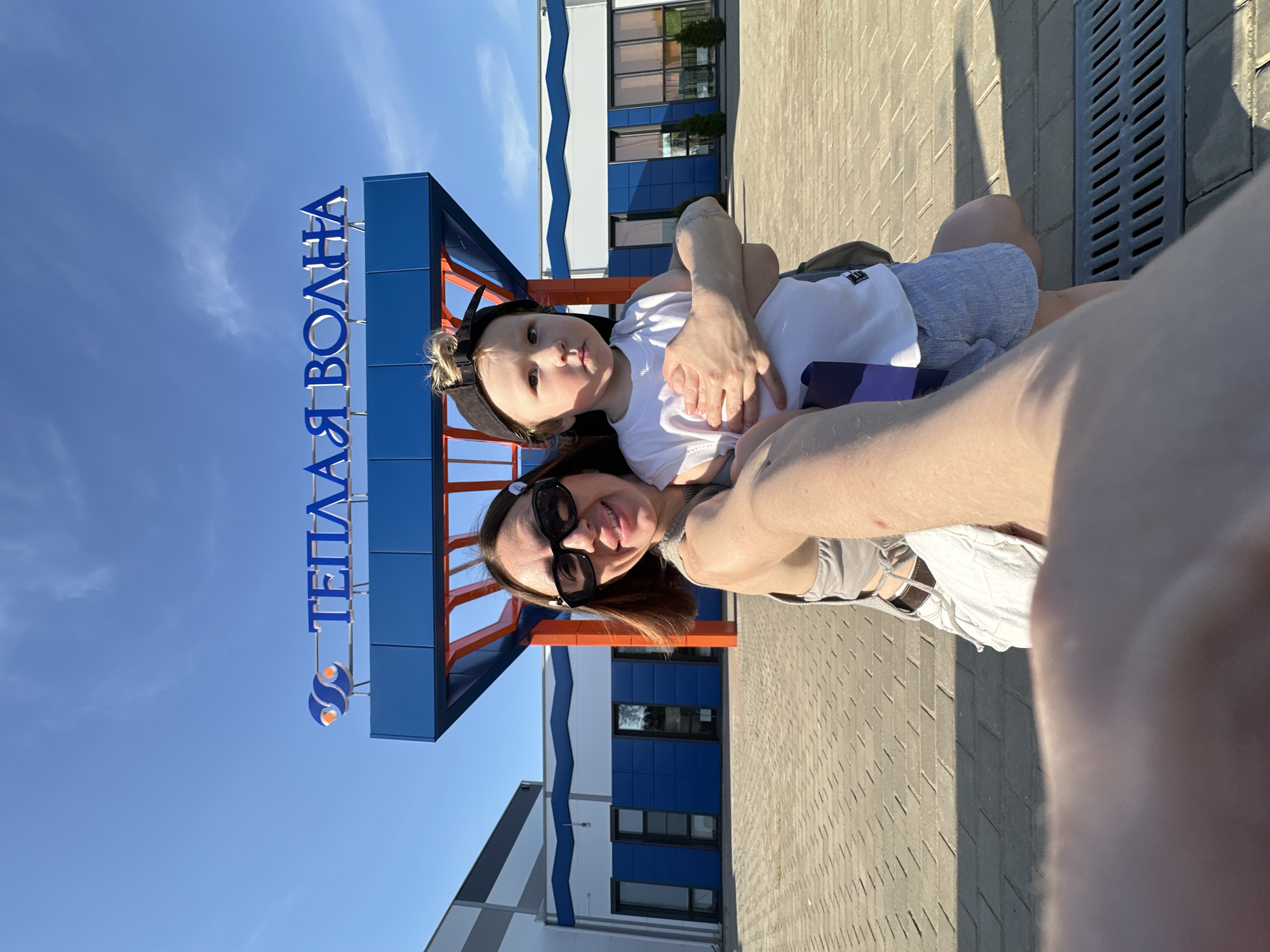 The image shows a woman and a young child in front of a building in Votkinsk, Russia. They appear to be taking a selfie, as the woman is holding the camera at arm's length. The child is wearing a white shirt and shorts, and a black baseball cap. The woman is wearing sunglasses. Behind them, there is a sign that reads "Теплая Волна" (Warm Wave). The building has a white and blue facade. The sky is bright blue. The scene is set outdoors on a paved area.