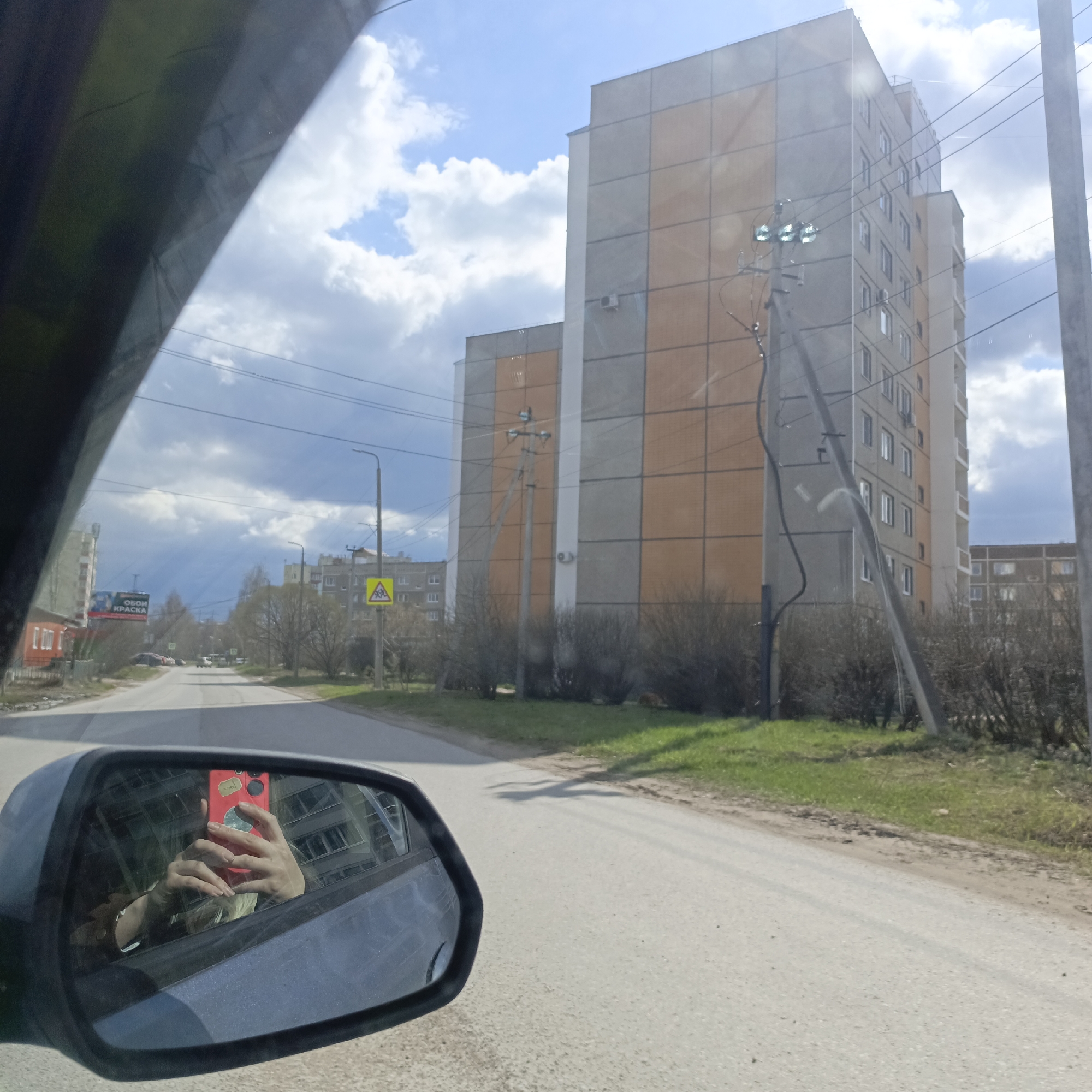 A view from inside a car driving on a paved road in what appears to be a residential area. The sky is blue with scattered clouds. Several multi-story buildings are visible in the background, with a combination of gray and orange panels on their facades. Power lines run across the street. The car's side mirror reflects the hands of a person holding a red smartphone, presumably taking the photograph. The location is the city of Unknown, Unknown.