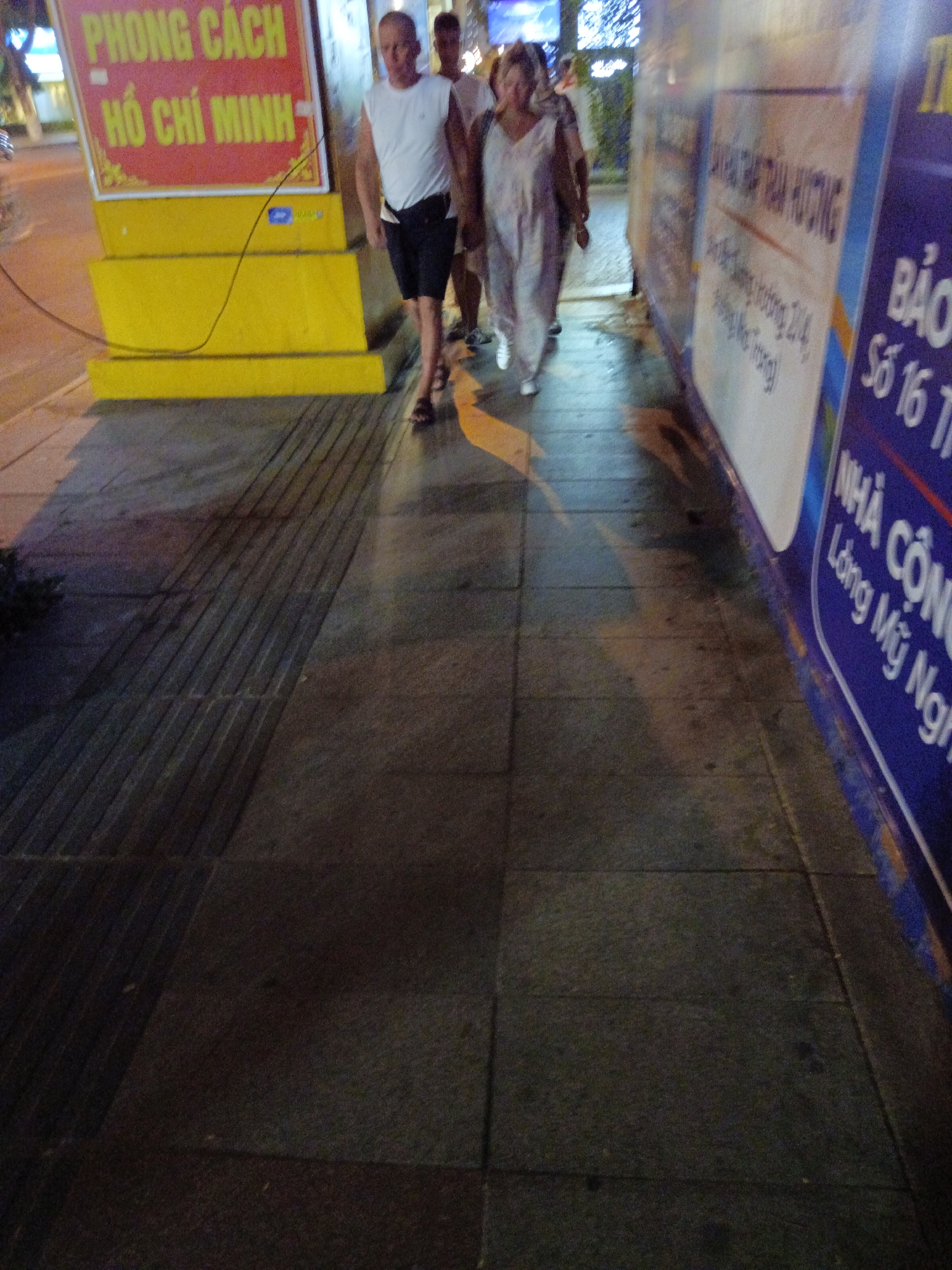 The image shows a street scene in Nha Trang, Vietnam at night. The foreground features a textured sidewalk with a pattern of raised lines. To the left, a large yellow structure displays text in Vietnamese, including "PHONG CÁCH" and "HỒ CHÍ MINH". In the mid-ground, three people are walking. A man in a white sleeveless shirt and dark shorts is in the center, looking ahead. To his right, a woman in a light-colored patterned dress is walking. Another person, partially obscured, is to the left of the man. In the background, a brightly lit sign and some foliage are visible. A blue and white sign with Vietnamese text is visible on the right side of the image, partially cut off. The overall lighting suggests artificial streetlights and illuminated signs.