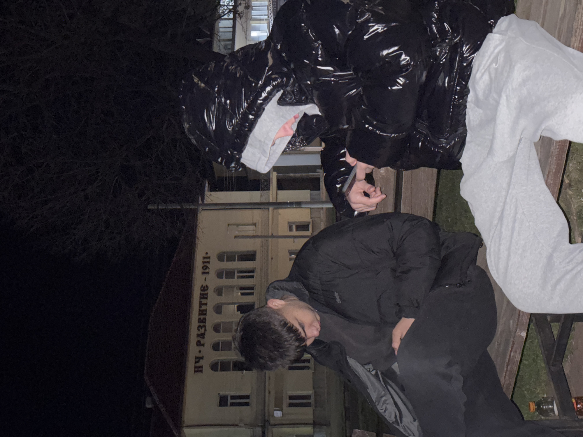 Two people are on a bench outside a building in Lehchevo, Bulgaria. The building has a sign in Cyrillic reading "НЧ. РАЗВИТИЕ-1911.". One person is wearing a shiny black hooded jacket, grey pants, and is holding a phone. The other person is wearing a black hooded jacket and black pants. It is dark outside. There is a tree to the left of the building.